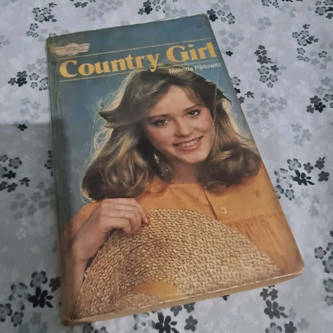 Country Girl 桌布 壁紙 — 復古書封面，一位微笑的女性在花卉圖案表面上拿著編織帽。
