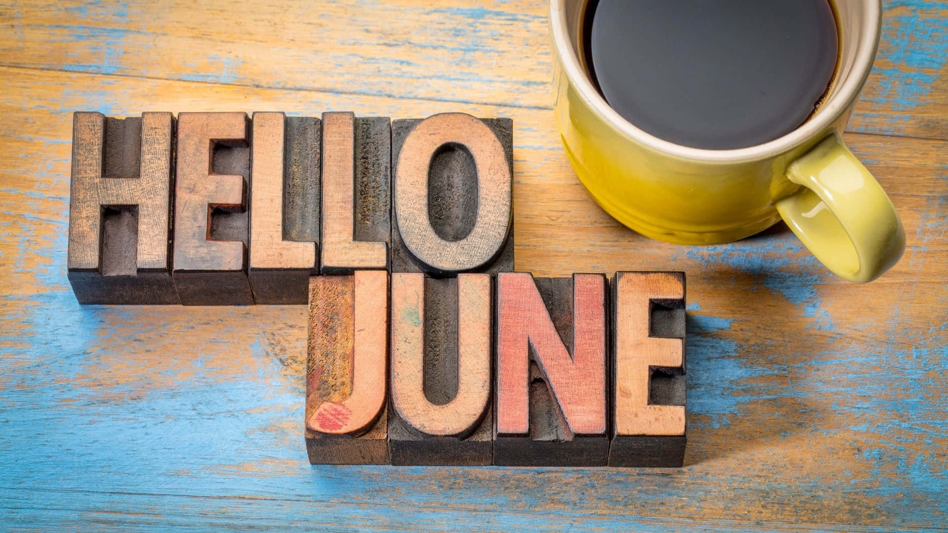 June 桌布 壁紙 — 復古木活字塊拼出哈囉六月，旁邊有一個黃色咖啡杯在風化的藍色木質表面上。