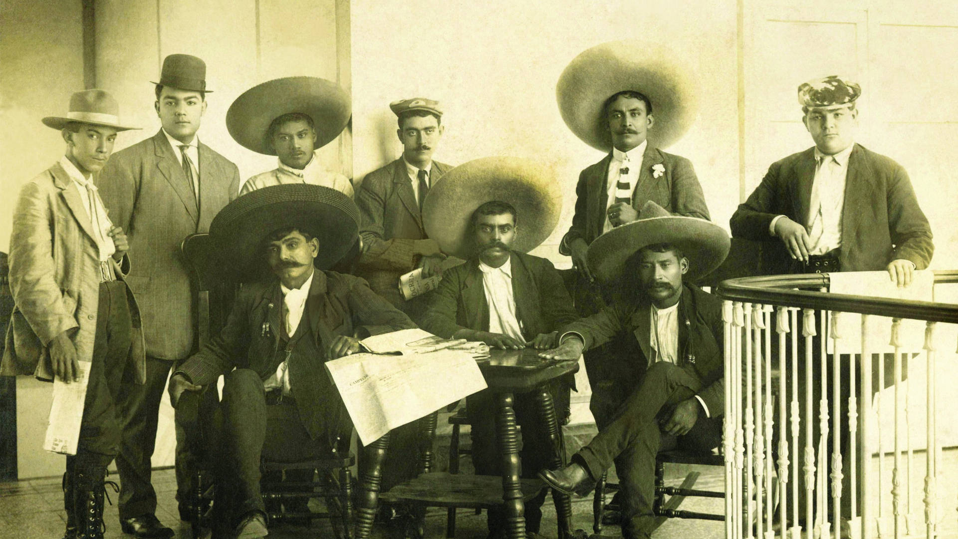 Mexican 桌布 壁紙 — 復古棕褐色調的八位男士穿著西裝和寬邊帽在室內欄杆旁正式擺姿勢的團體肖像。