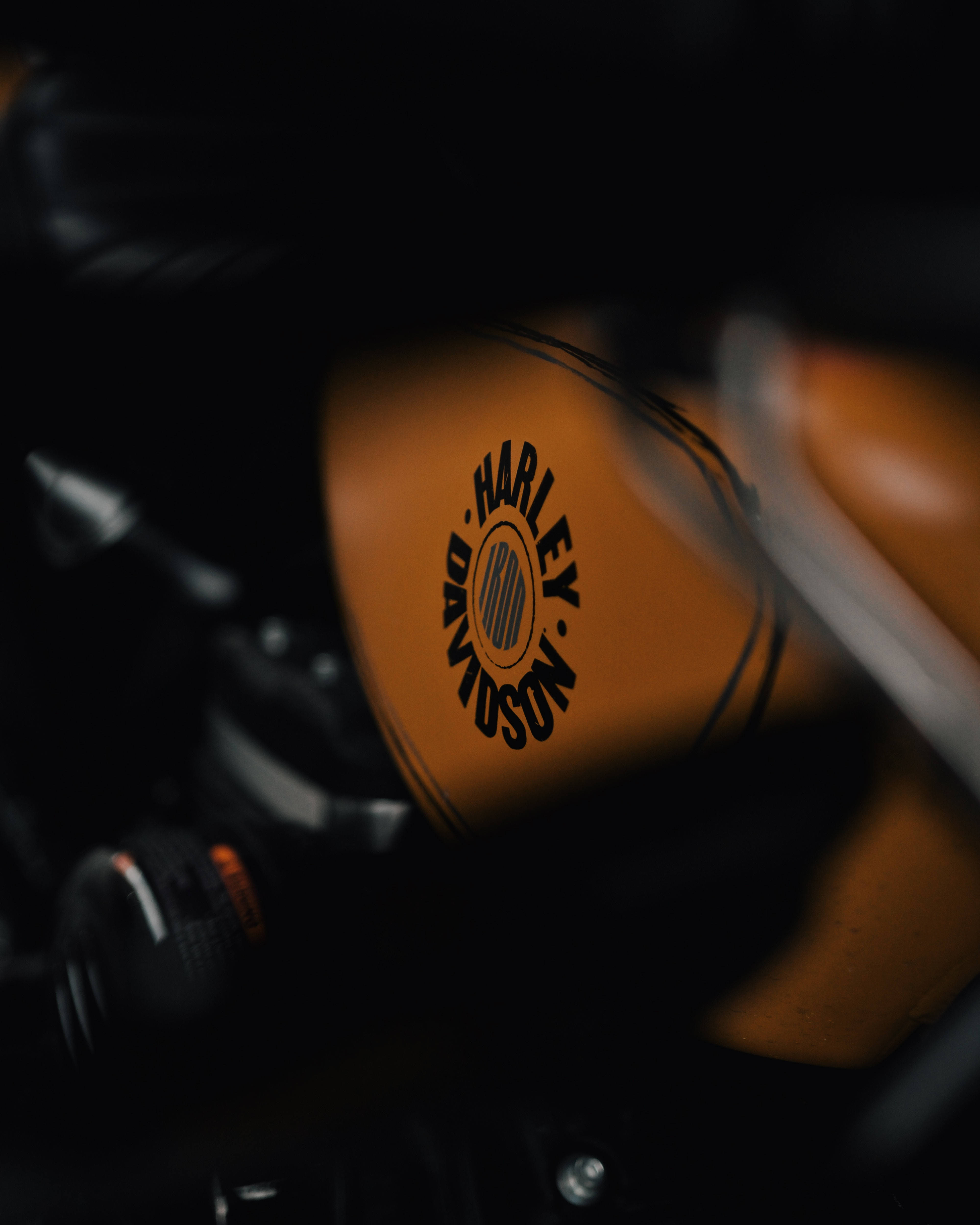 Harley 桌布 壁紙 — 復古橙色Harley-Davidson機車油箱特寫，黑色標誌，背景為模糊的深色機車零件。