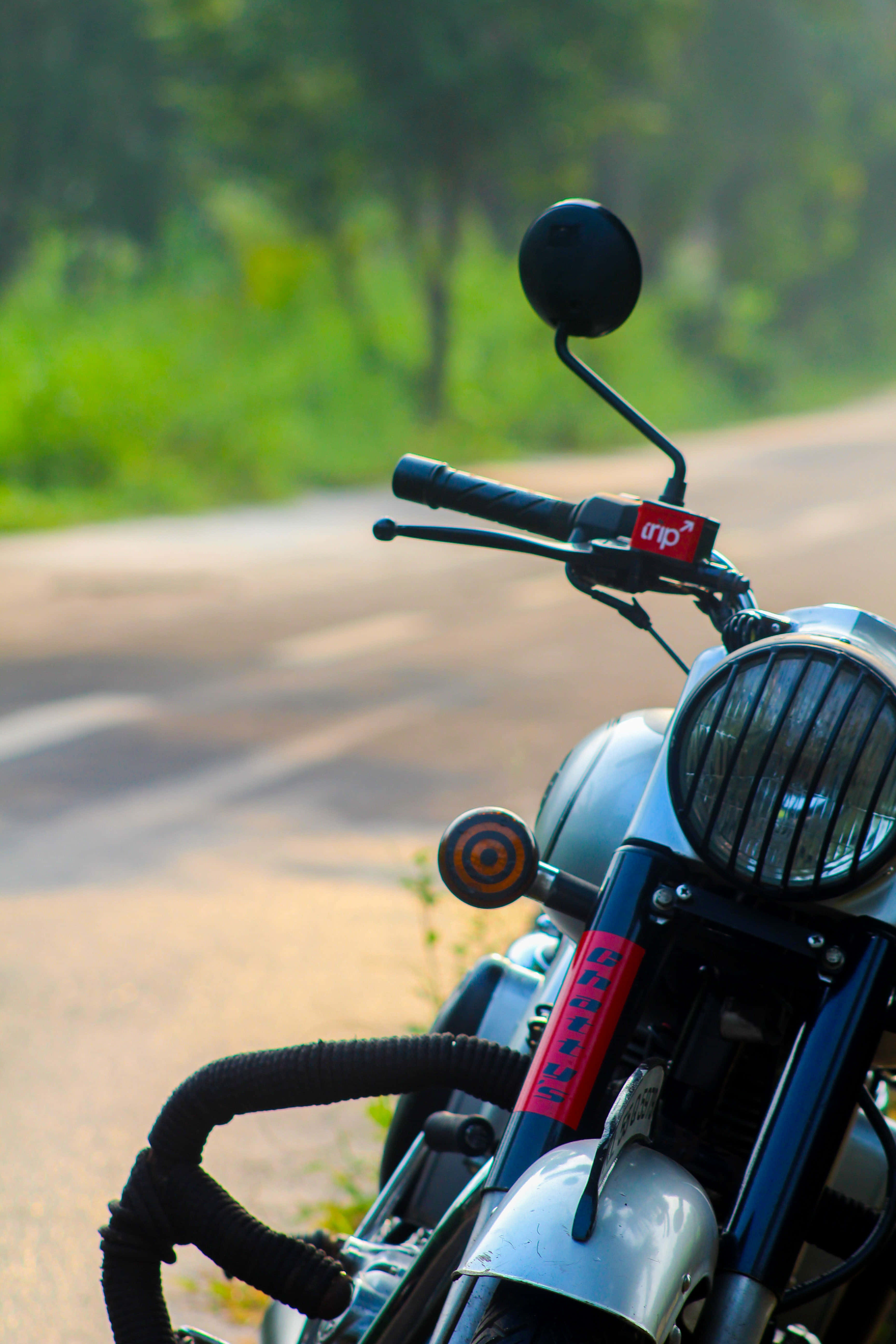 Royal Enfield Bullet 桌布 壁紙 — 復古機車的把手和頭燈特寫，背景是陽光明媚的鄉村道路和模糊的綠植。