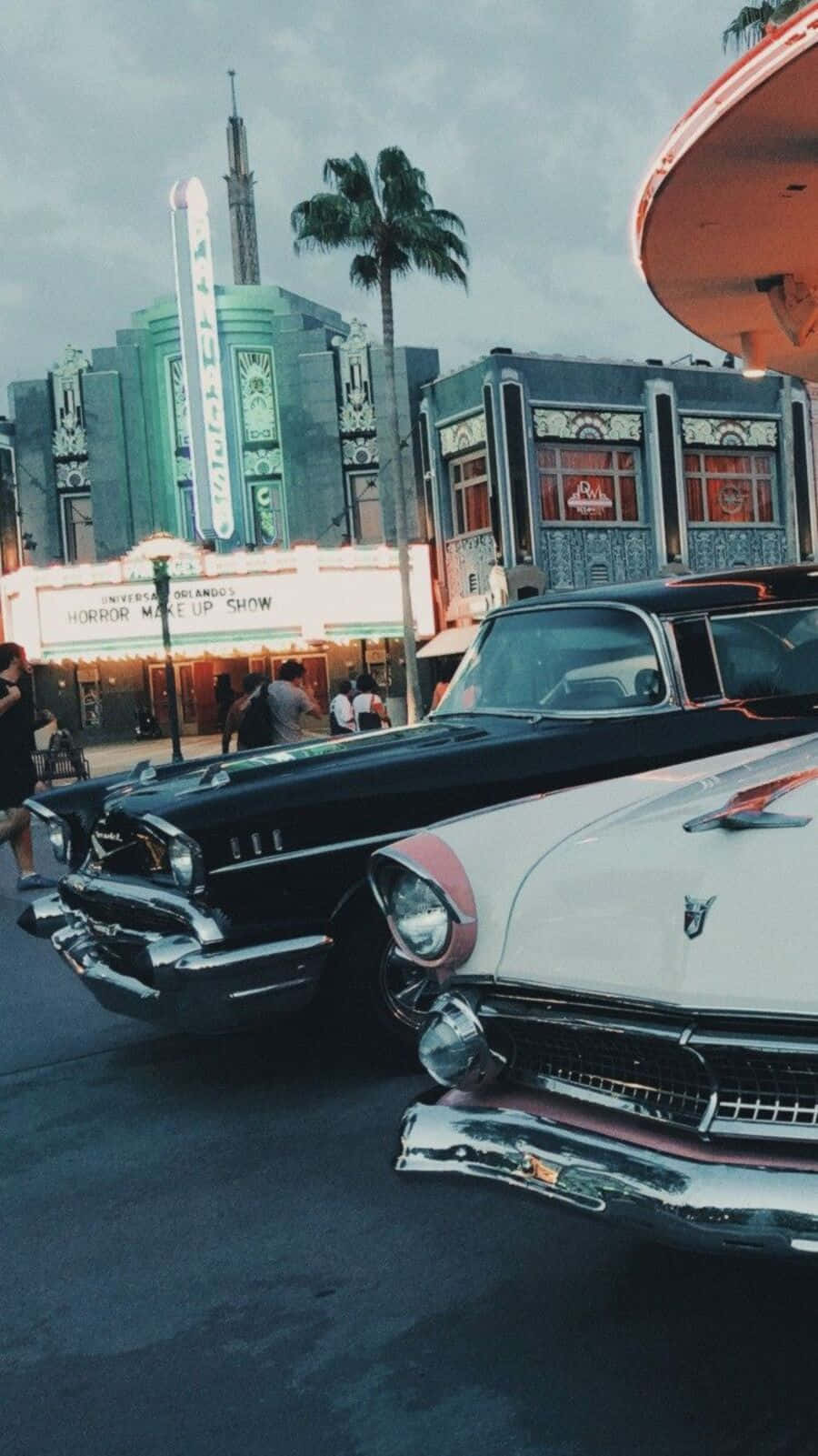 90's Vintage Retro Aesthetic 桌布 壁紙 — 復古汽車在暮色天空下，在棕櫚樹和柔和發光的霓虹燈招牌旁邊的復古劇院旁閃閃發光。