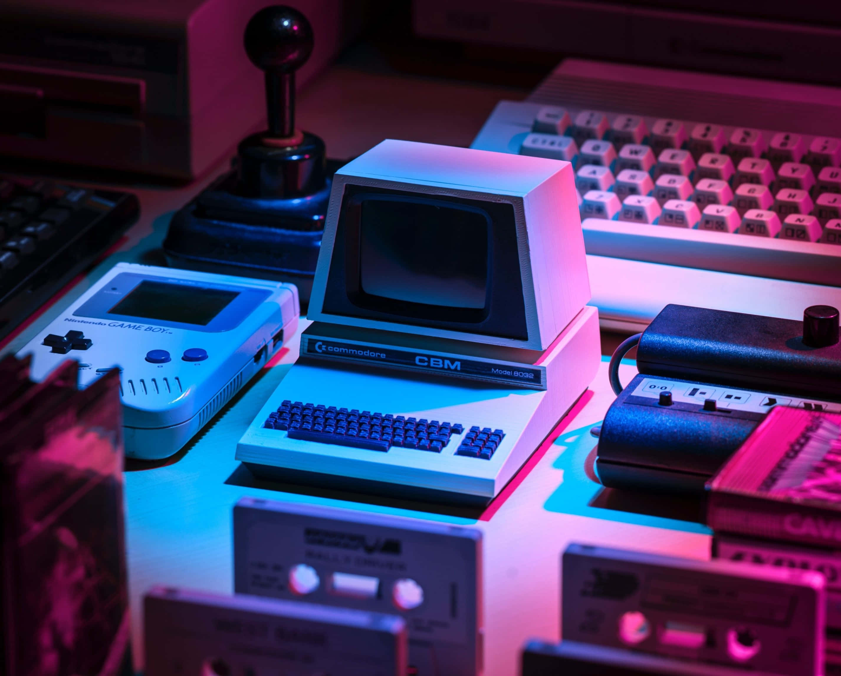 Retro Aesthetic 桌布 壁紙 — 復古遊戲和電腦設備在雜亂的桌子上沐浴在霓虹粉紅和藍光中。