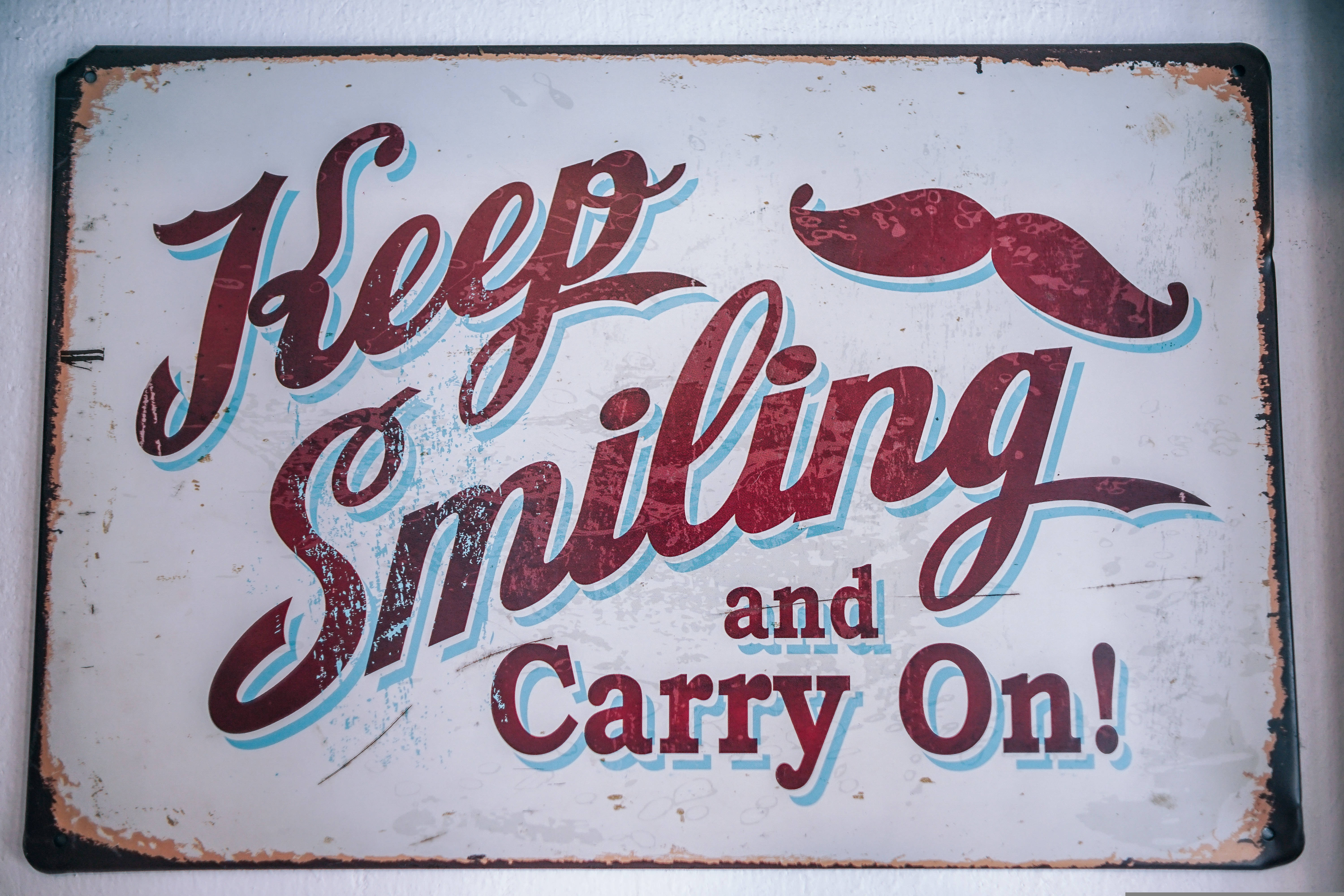 Inspirational Quotes 桌布 壁紙 — 復古風化標誌，紅色草書文字寫著「Keep Smiling and Carry On!」，頂部有紅色鬍子。