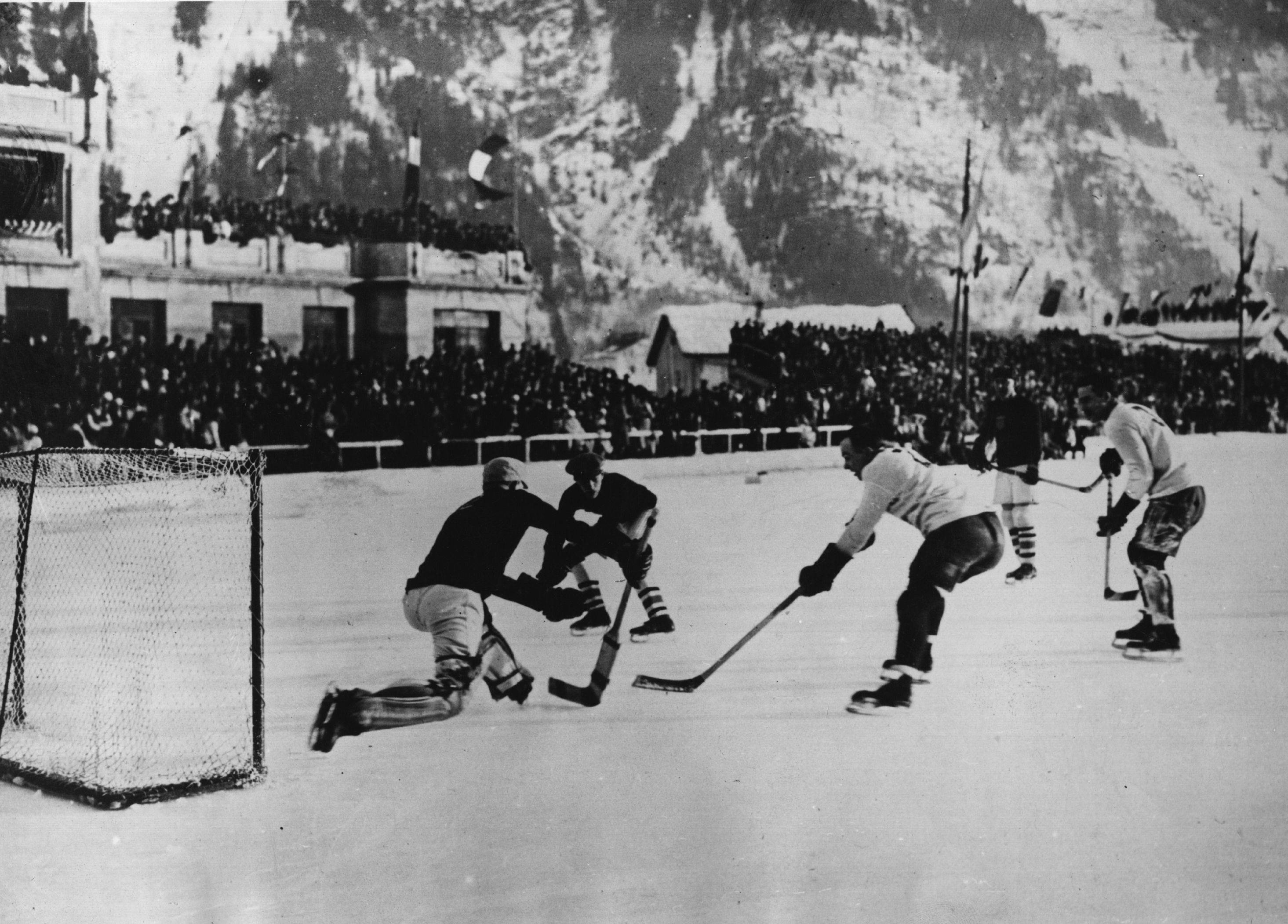 Winter Olympics 桌布 壁紙 — 復古黑白照片，冰球運動員在雪地溜冰場上激烈競爭，觀眾在觀眾席觀看，背景是山脈。
