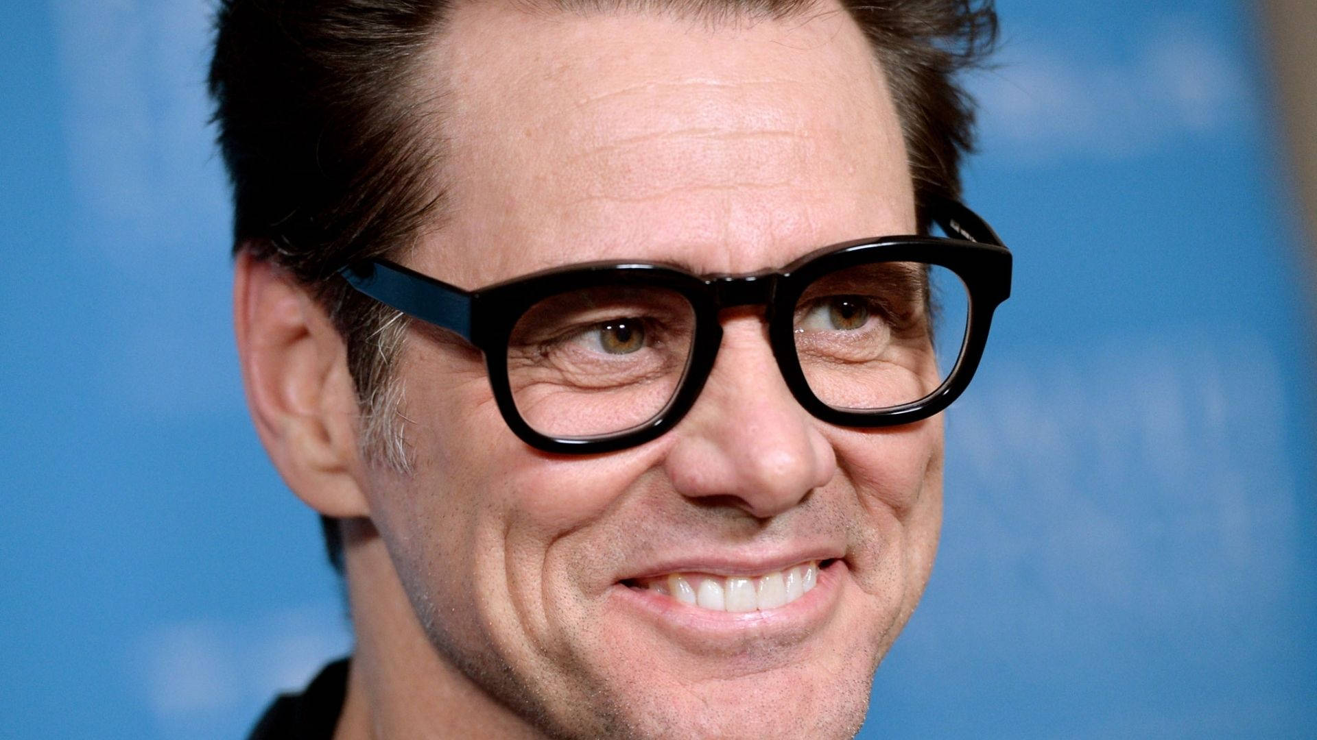 Jim Carrey 桌布 壁紙 — 微笑男士戴粗黑框眼鏡的特寫肖像，背景是柔和藍色。