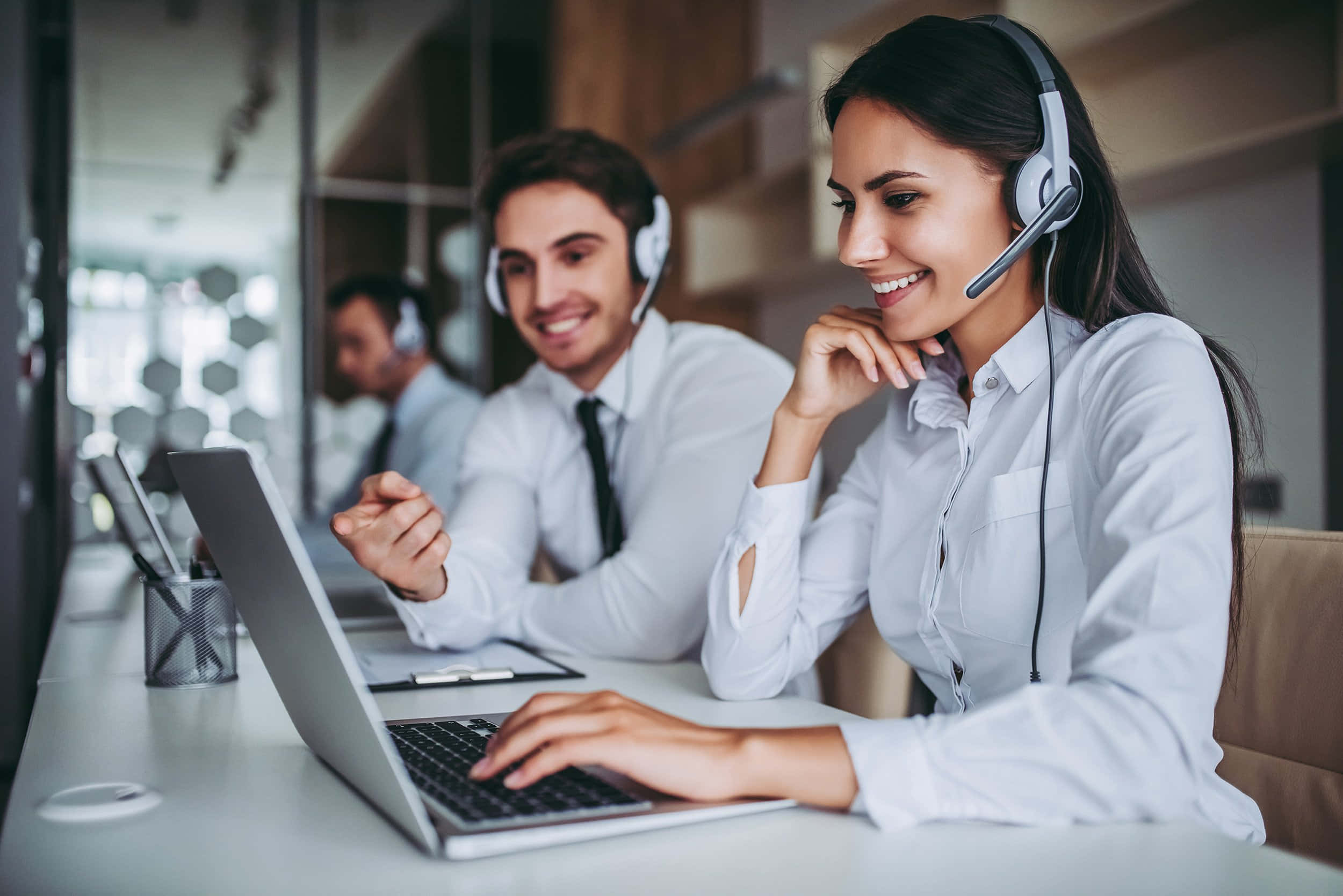 Telemarketing 桌布 壁紙 — 微笑的客戶服務人員戴著耳機，在設有玻璃隔間和溫暖照明的現代辦公室中使用筆記型電腦工作。