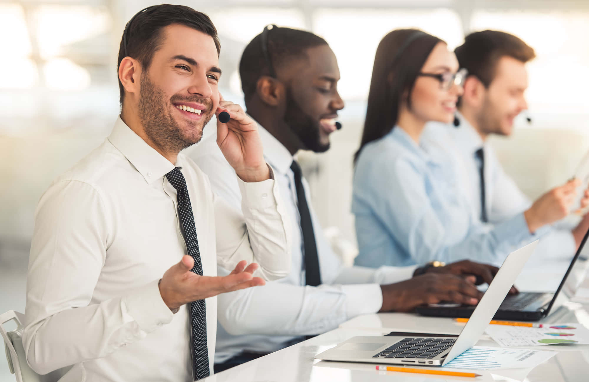 Telemarketing 桌布 壁紙 — 微笑的客戶服務人員穿著白襯衫和領帶，在明亮現代化的客服中心使用筆記型電腦工作。