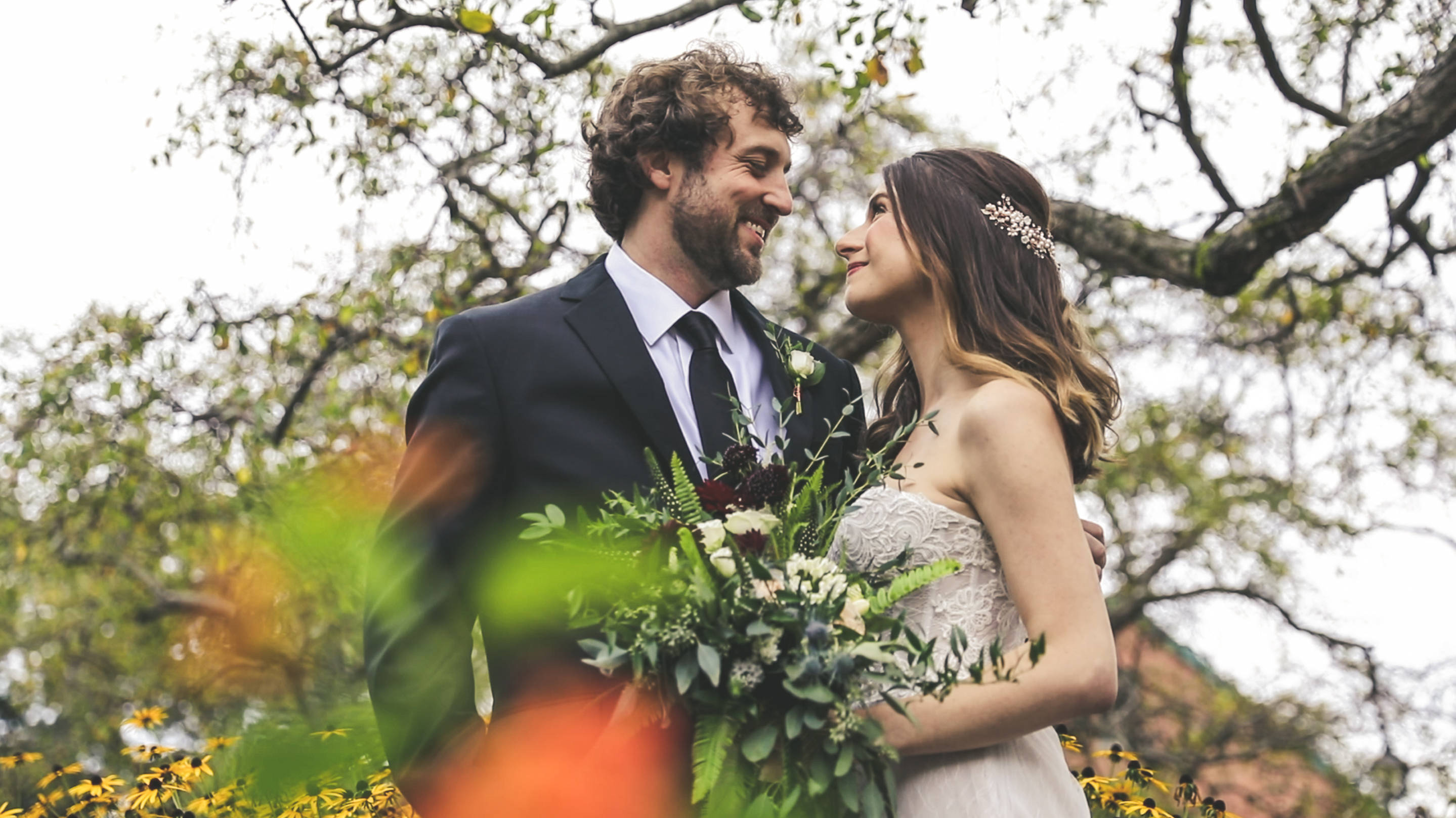 Groom 桌布 壁紙 — 微笑的新郎和新娘在枝葉茂密的樹下深情對望，前景有模糊的黃色花朵。