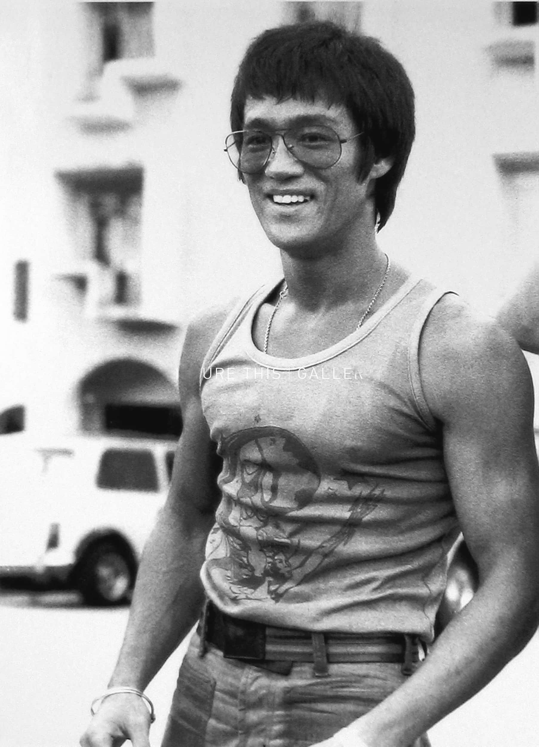 李小龍 桌布 壁紙 — 微笑的李小龍身穿背心和眼鏡，自信地站在模糊的城市背景前。