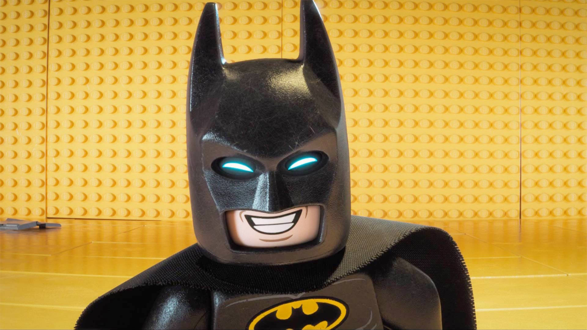 Batman Animated 桌布 壁紙 — 微笑的樂高蝙蝠俠人偶特寫，藍色眼睛發光，背景為黃色磚牆。