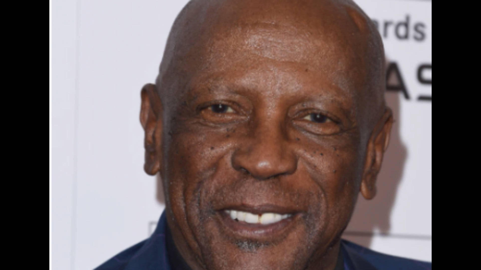 Celebrity 桌布 壁紙 — 微笑的禿頭黑人男子特寫肖像，眼神溫暖，身穿藍色襯衫，背景為白色活動背景。
