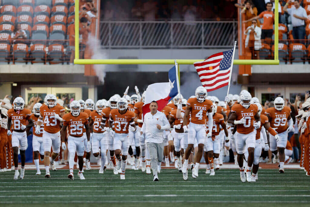 Texas Football 桌布 壁紙 — 德州足球隊身穿橙色球衣，在球場燈光下高舉美國國旗和德州州旗跑入球場。