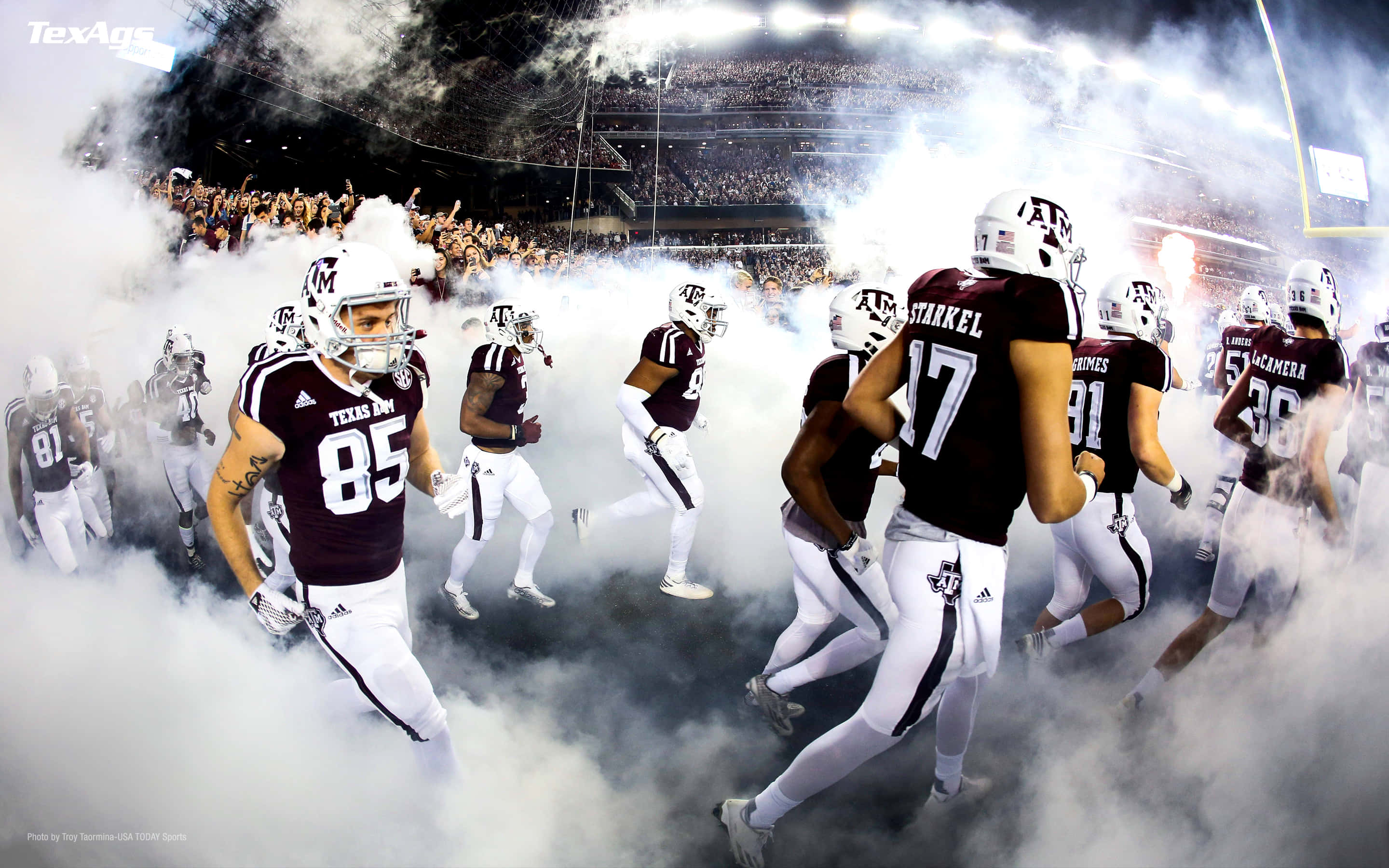 Texas Football 桌布 壁紙 — 德州農工大學美式足球員衝破煙霧進入球場，球迷在體育館燈光下從看台歡呼。