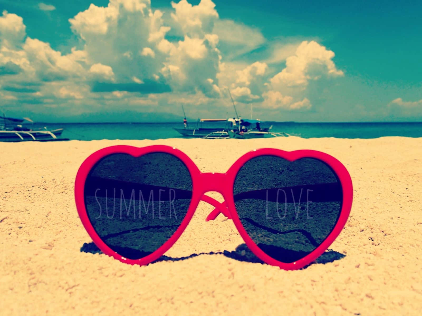 海灘 桌布 壁紙 — 心形粉紅色太陽眼鏡，上面有'SUMMER'和'LOVE'字樣，放置在沙灘上，背景有船隻和多雲的天空。