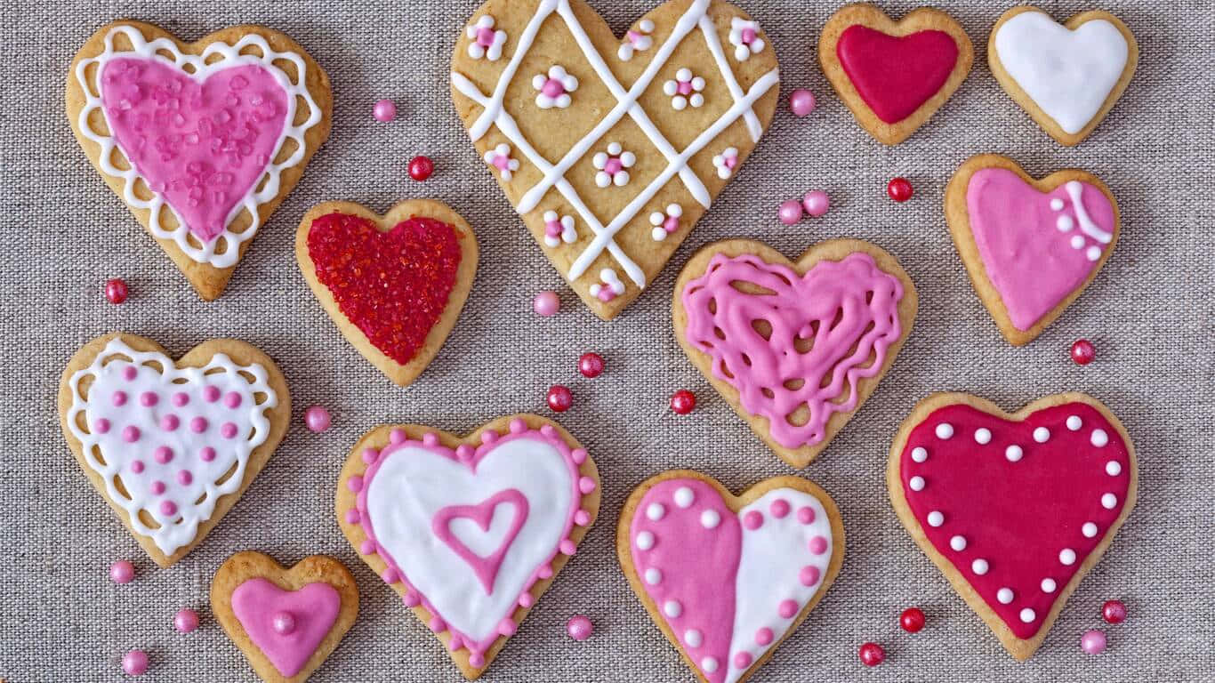 1366x768 Cookies 桌布 壁紙 — 心形餅乾，裝飾有粉紅、白色和紅色糖霜及糖粒，排列在帶紋理的米色表面上。