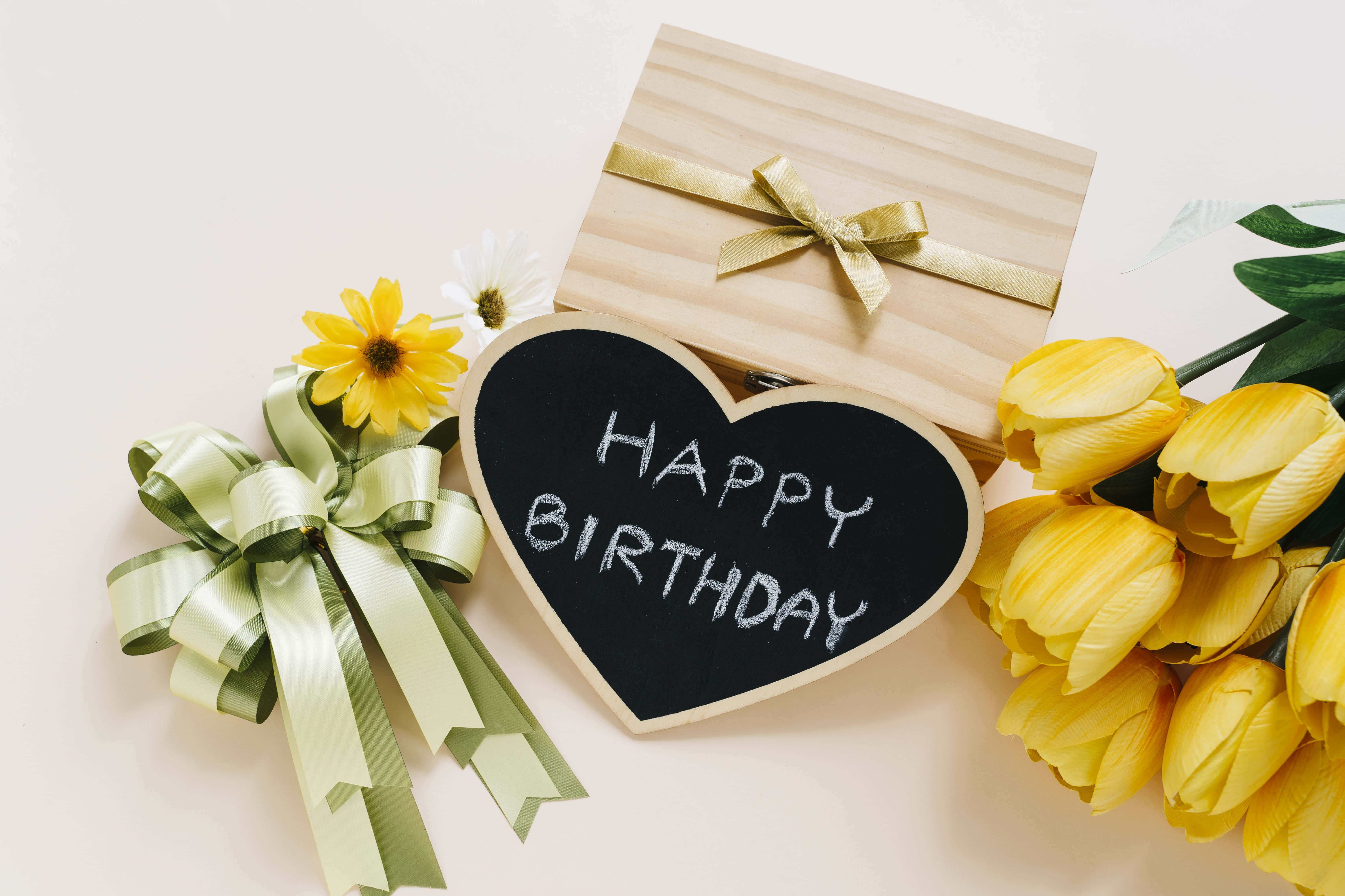 生日快樂 桌布 壁紙 — 心形黑板寫著「生日快樂」，周圍有黃色鬱金香、綠色蝴蝶結和木製禮物盒，放在白色表面上。