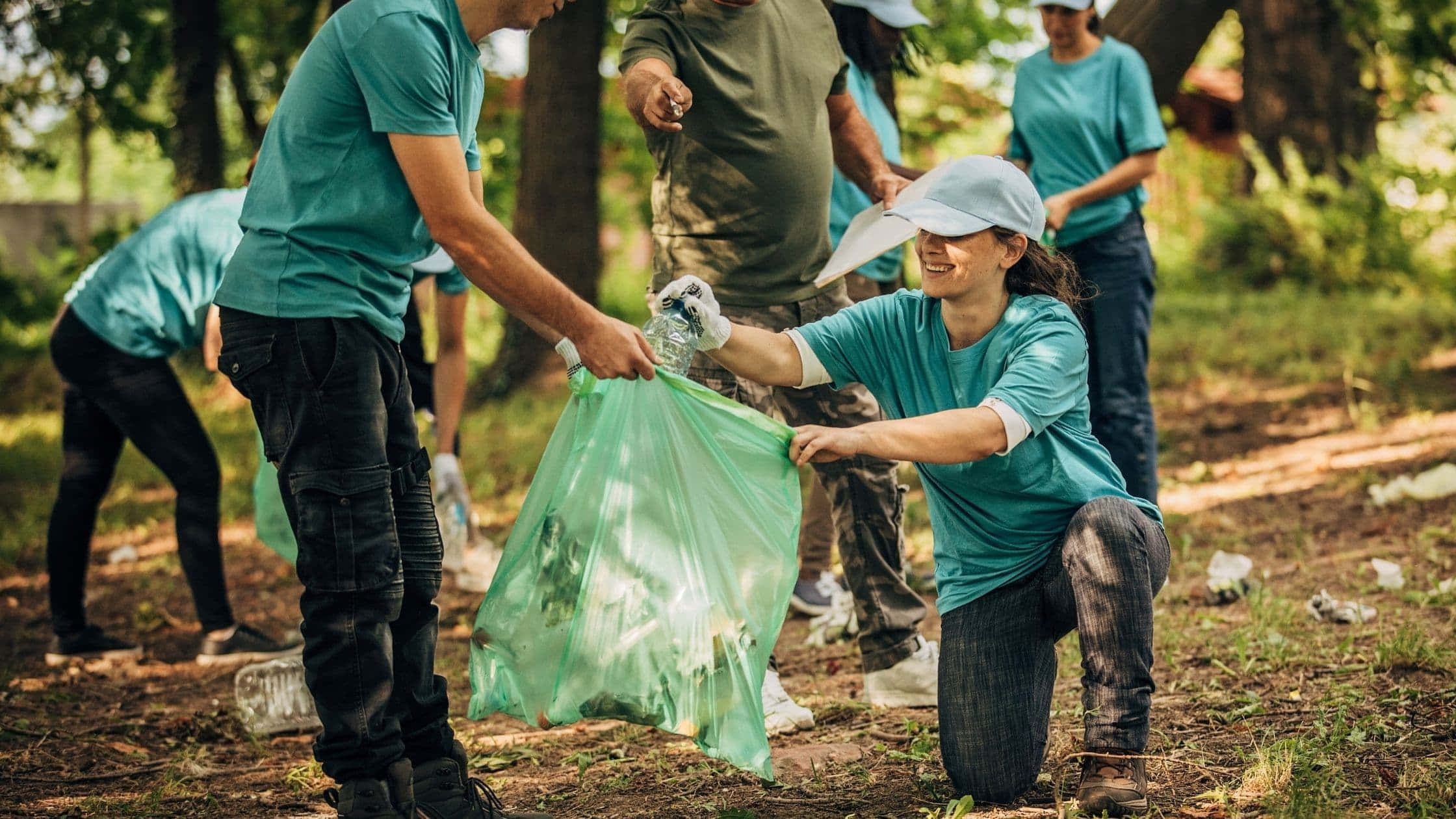 environment 桌布 壁紙 — 志工穿著青綠色T恤在明亮的日光下於森林空地收集垃圾。