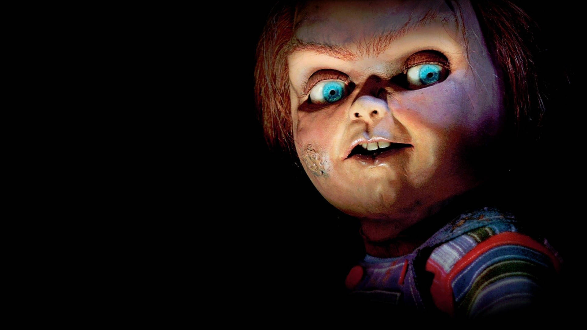 Chucky 桌布 壁紙 — 恐怖娃娃的特寫，擁有發光的藍色眼睛和邪惡的笑容，背景為黑色。