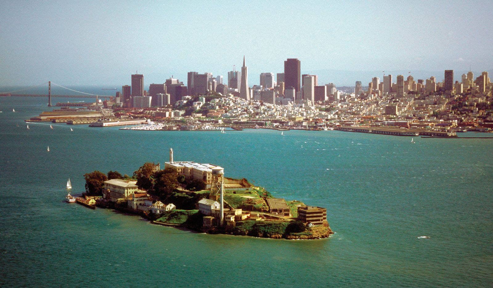 城市 桌布 壁紙 — 惡魔島上的歷史建築坐落在碧綠的海灣中，背景是舊金山的天際線和金門大橋。