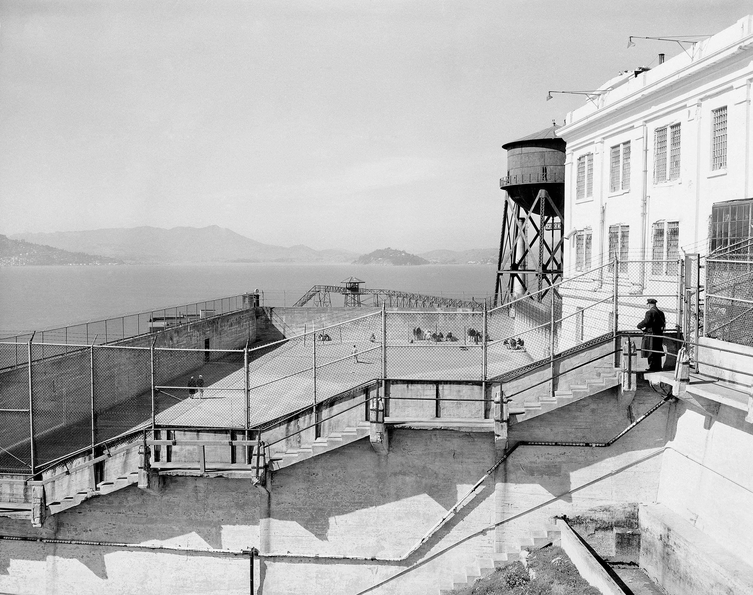 Prison 桌布 壁紙 — 惡魔島監獄的黑白照片，有圍欄庭院、海洋和遠處山丘，天空晴朗。