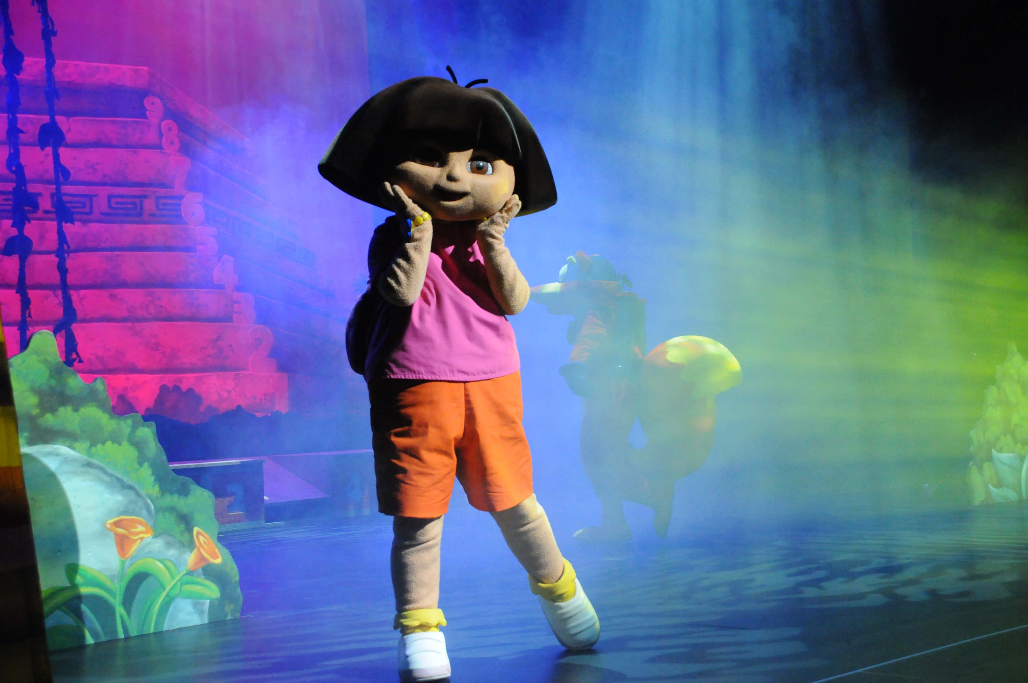 Funny Dora 桌布 壁紙 — 愛探險的Dora吉祥物在一個色彩繽紛的舞台上跳舞，舞台被鮮豔的紫色、綠色和藍色聚光燈照亮。