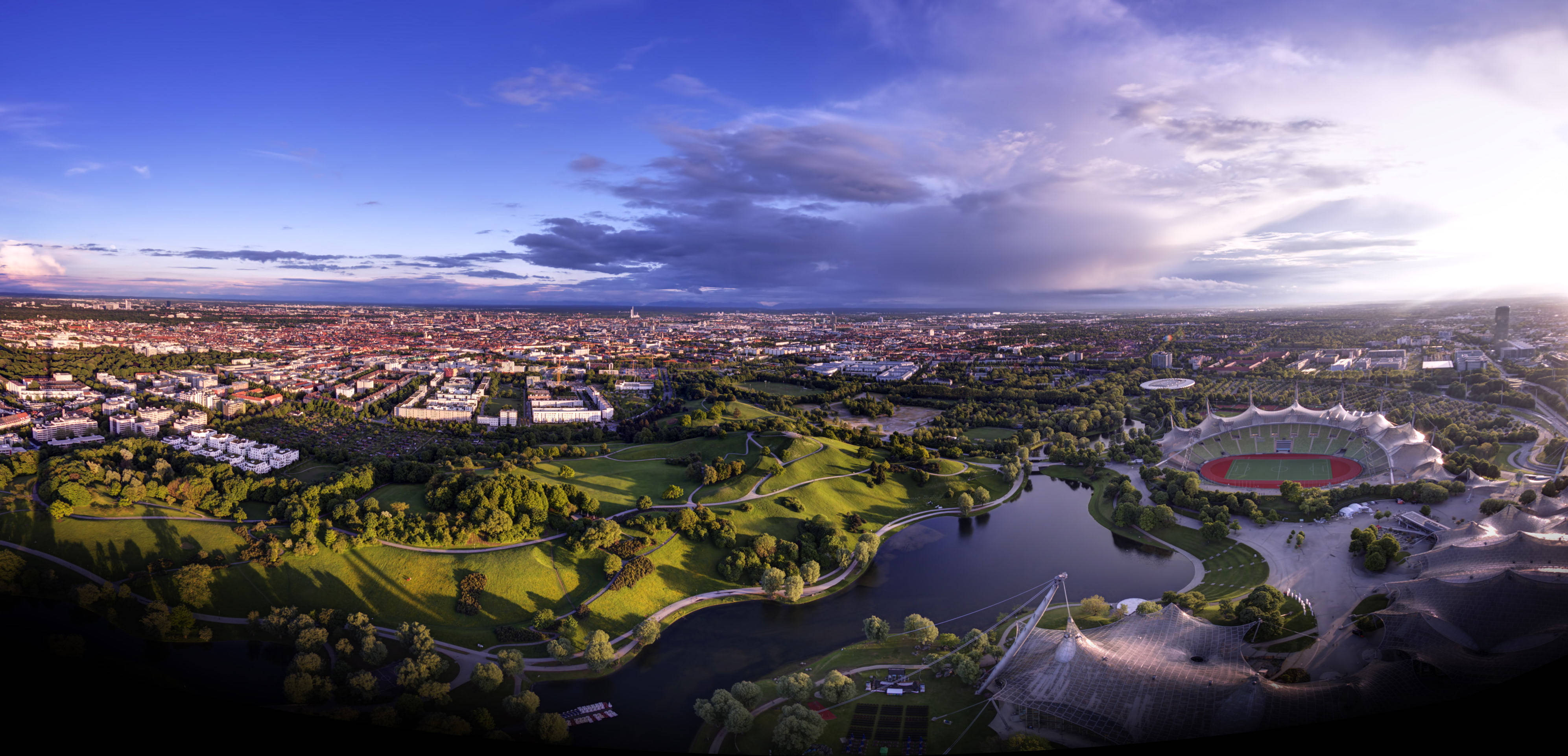 Munich 桌布 壁紙 — 慕尼黑奧林匹克公園的鳥瞰圖，夕陽西下，綠草如茵，河流蜿蜒，標誌性體育場在戲劇性的天空下。