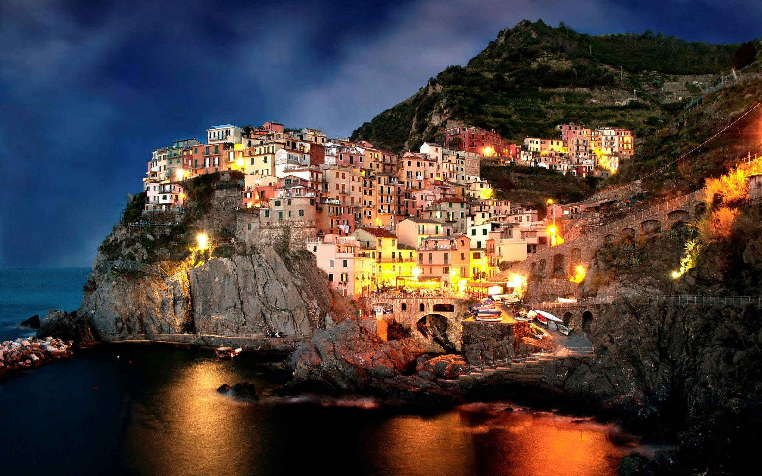 Amalfi Coast 桌布 壁紙 — 懸崖邊的村莊在夜晚溫暖地發光，色彩繽紛的建築依偎在黑暗的岩石間，並在平靜的海面上倒映。