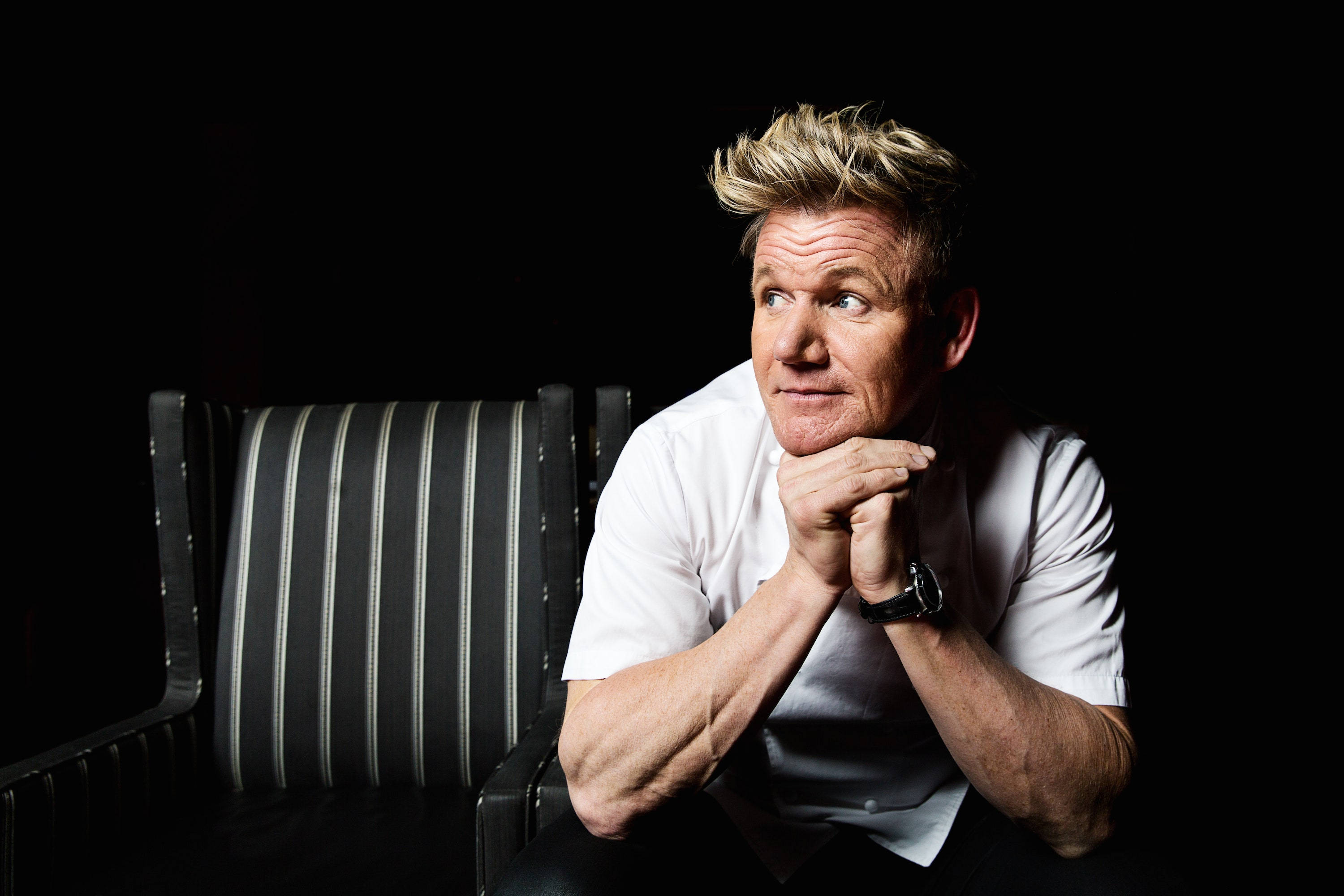 Gordon Ramsay 桌布 壁紙 — 戈登·拉姆齊坐在黑色背景前的條紋椅子上，雙手交叉放在下巴下，表情嚴肅地看向一旁。