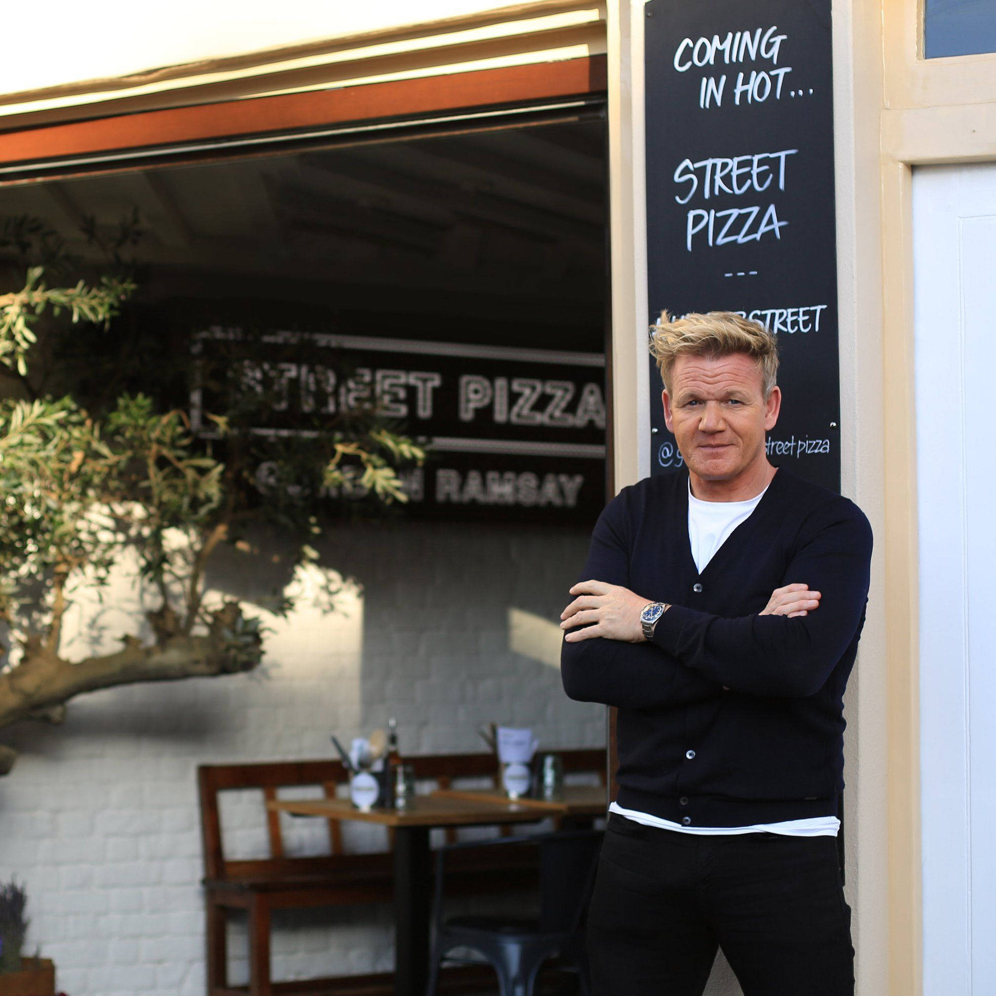 Gordon Ramsay 桌布 壁紙 — 戈登·拉姆齊自信地交叉雙臂，站在他的街頭披薩餐廳外，在明亮的日光下。