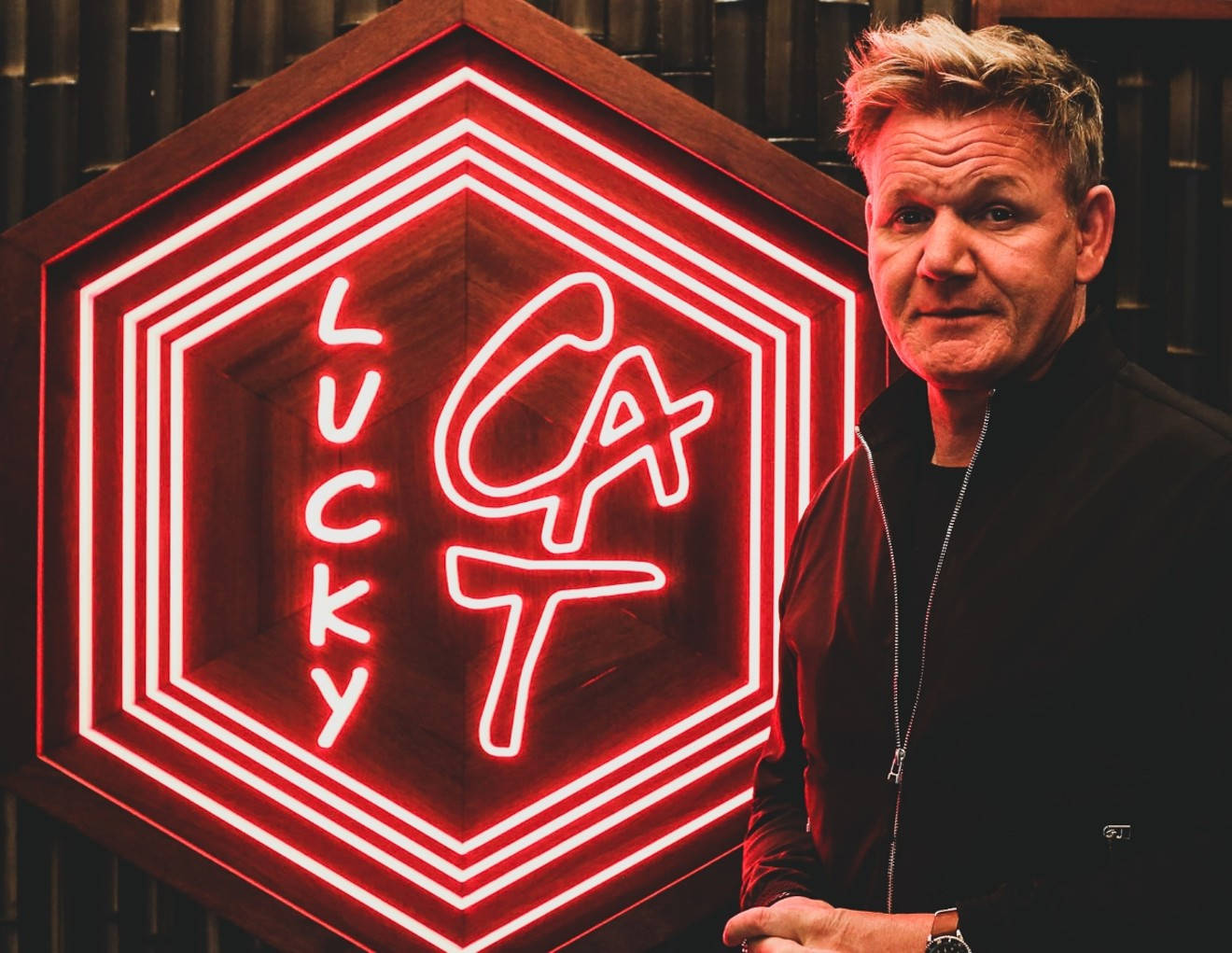 Gordon Ramsay 桌布 壁紙 — 戈登·拉姆齊自信地站在一個發光的紅色六角形霓虹燈招牌旁，招牌上寫著「Lucky Cat」，室內燈光昏暗。