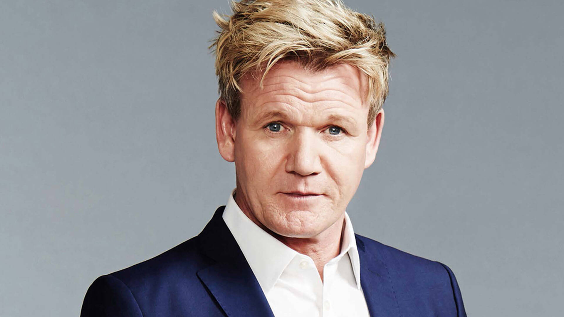 Gordon Ramsay 桌布 壁紙 — 戈登·拉姆齊身穿俐落的深藍色西裝和白色襯衫，在純灰色背景前，目光直接而強烈。