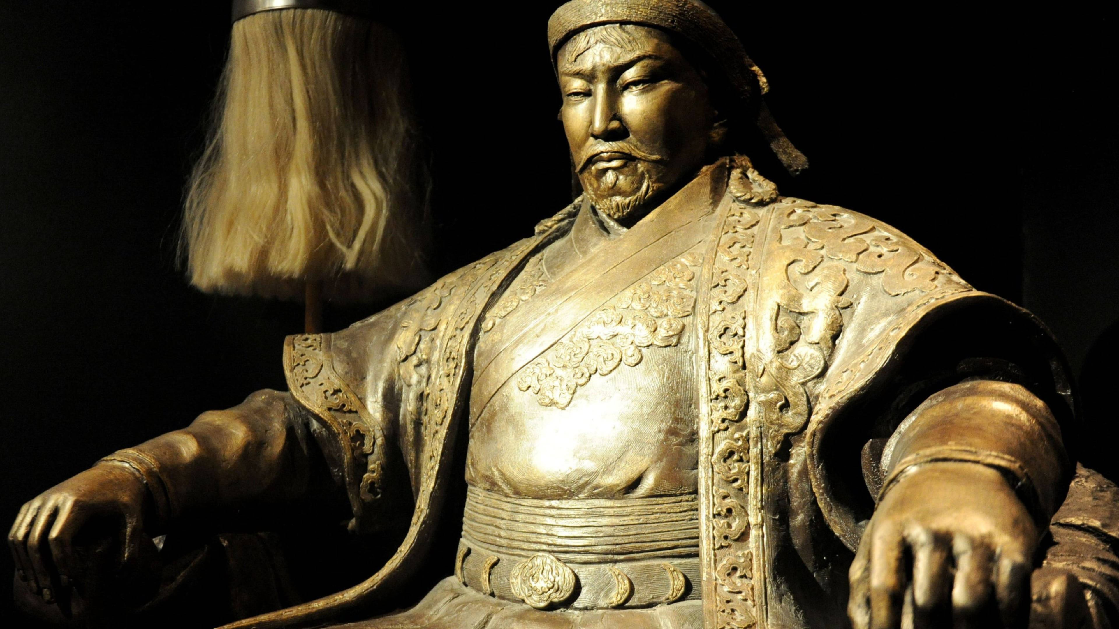 Mongolia 桌布 壁紙 — 成吉思汗的金色雕像，身穿華麗盔甲，頭戴禮儀帽，腰間飾有精緻腰帶，在戲劇性的聚光燈下威嚴端坐。