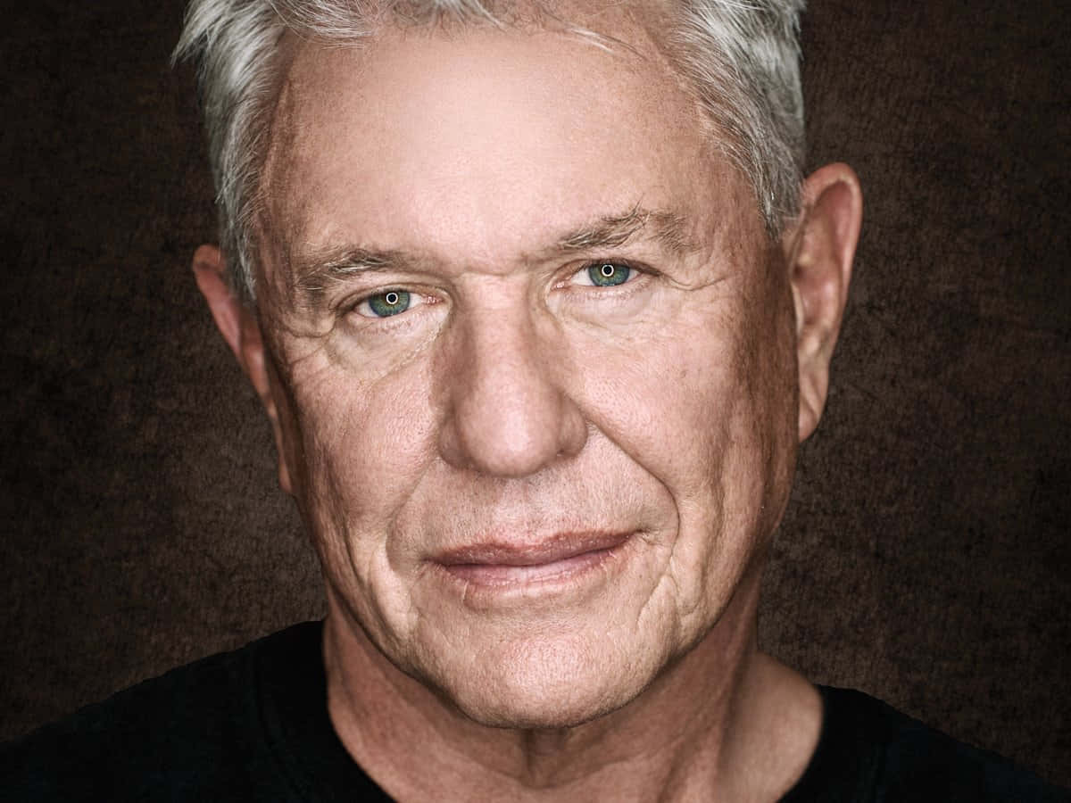 Tom Berenger 桌布 壁紙 — 成熟男士的頭像特寫，灰白頭髮，一雙強烈的藍眼睛，背景為深色紋理。
