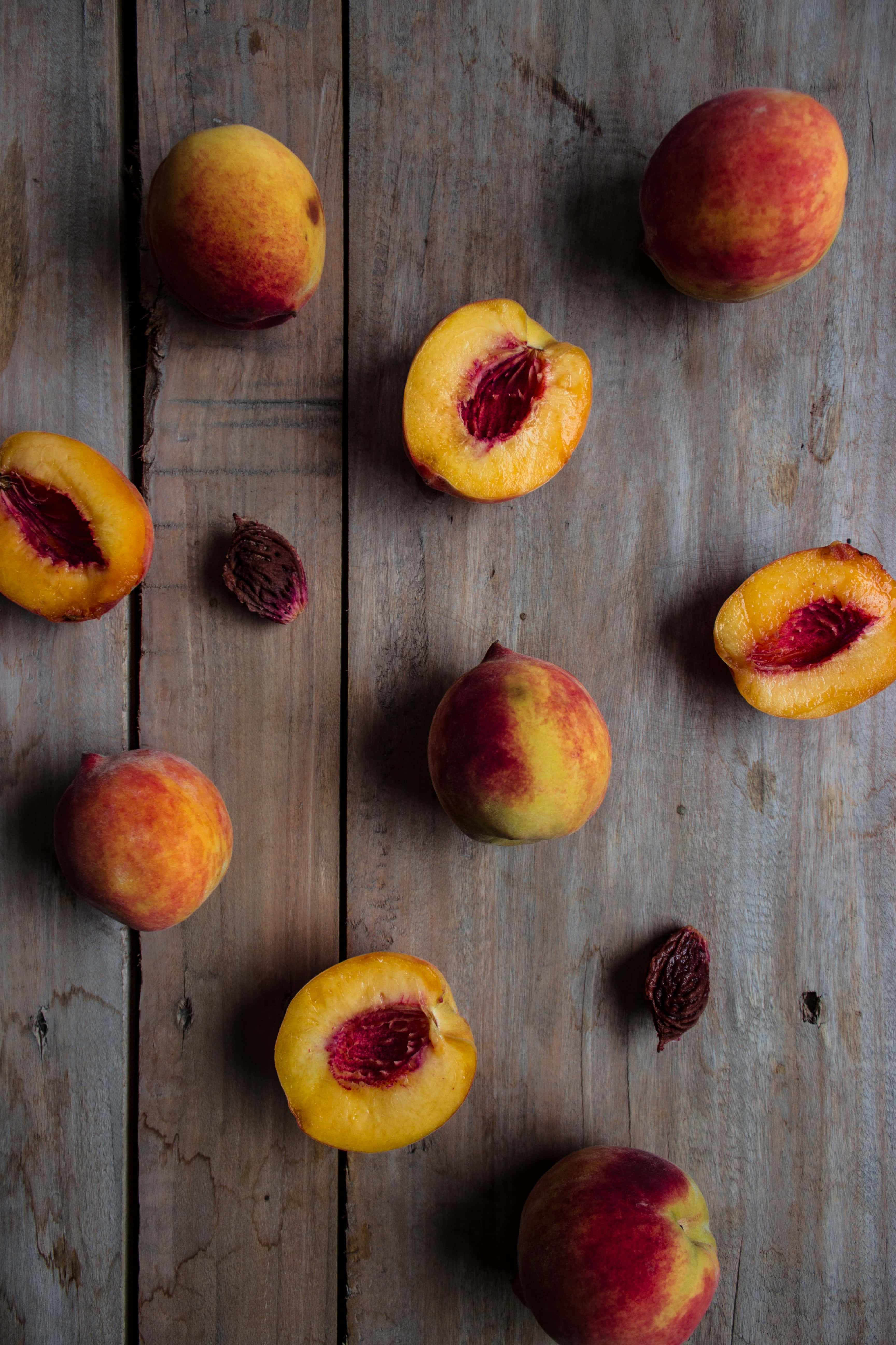 Peaches 桌布 壁紙 — 成熟的桃子，完整和切半，果核散落在風化的木質表面。