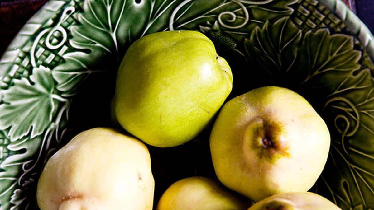 Quince 桌布 壁紙 — 成熟的榲桲放在一個帶有葉子浮雕圖案的深綠色陶瓷碗中，光線柔和。