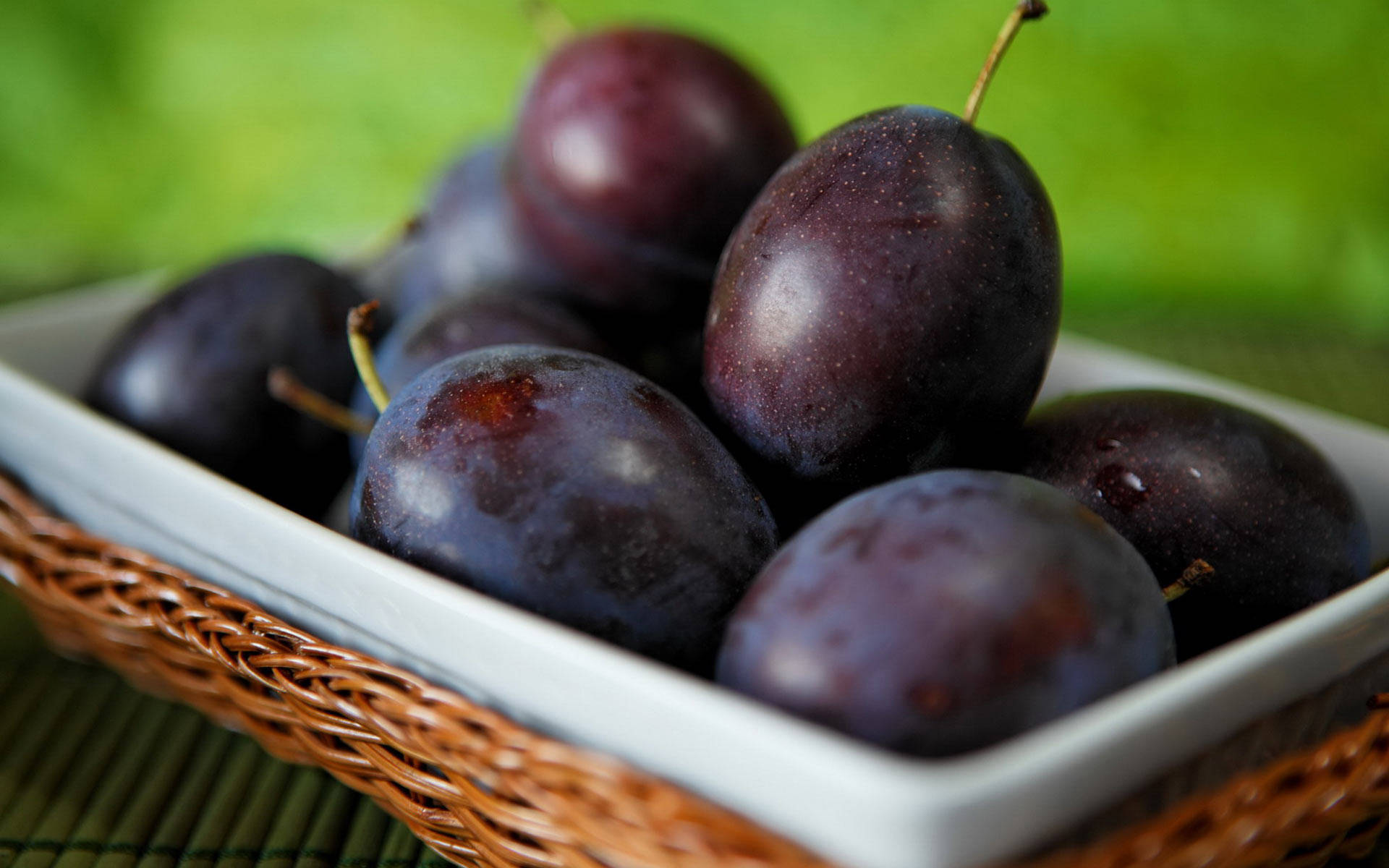 Fruits 桌布 壁紙 — 成熟的深紫色李子放在白色陶瓷碗中，碗置於編織柳條籃內，襯以柔和綠色背景，閃閃發光。