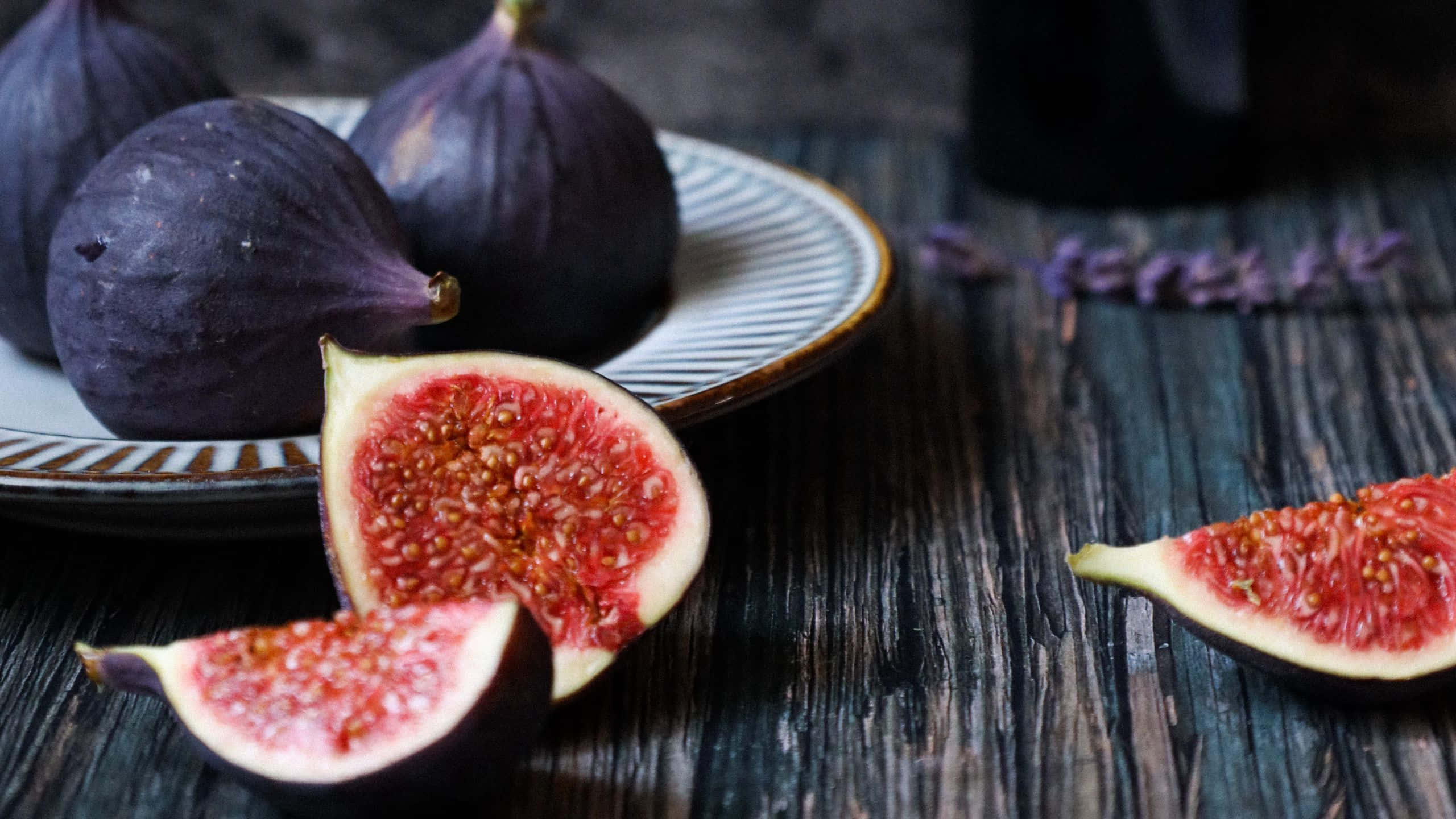 Purple Figs 桌布 壁紙 — 成熟的紫色無花果切開，露出多汁的紅色果肉，放在深色木桌上的一個條紋盤子上。