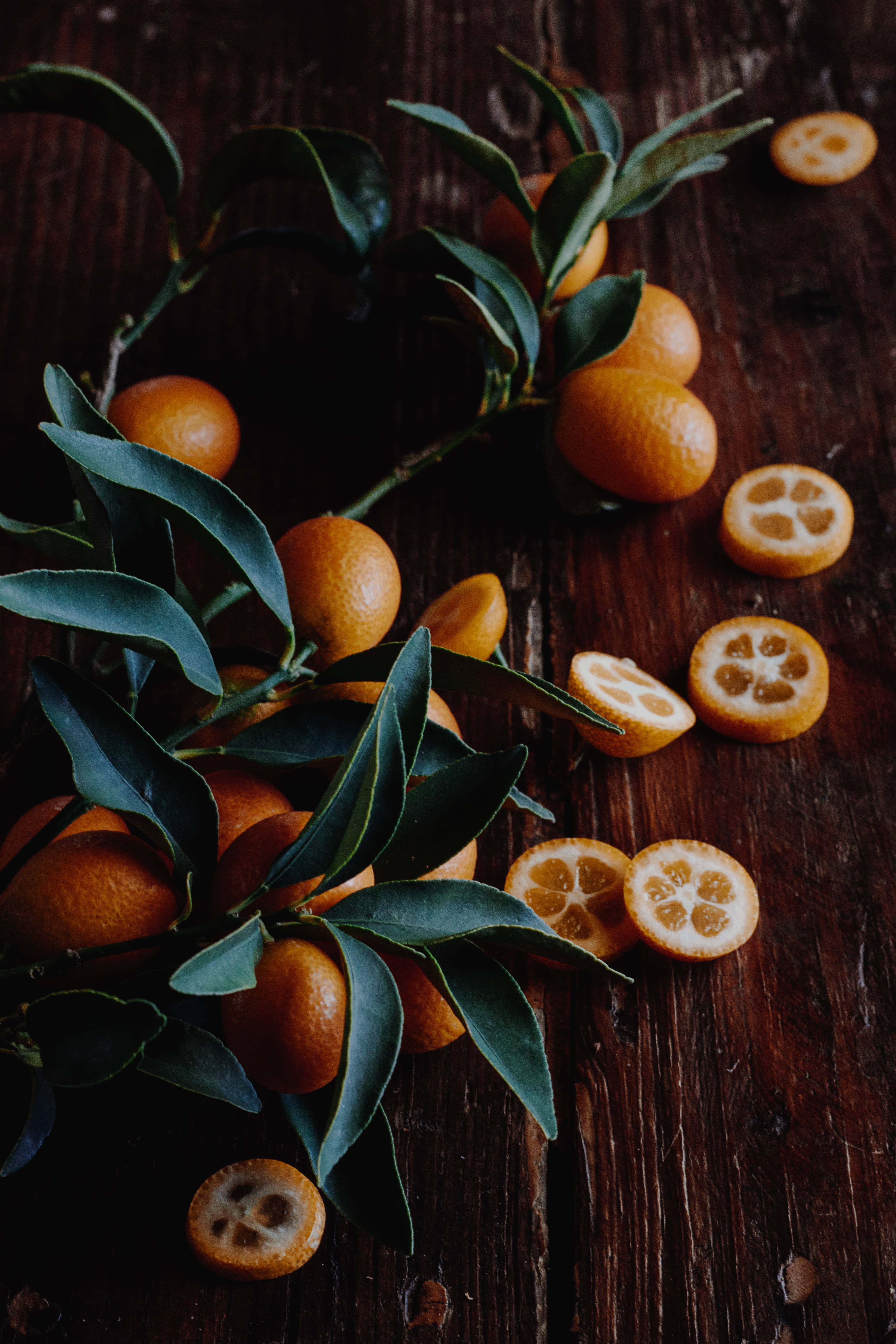 citrus 桌布 壁紙 — 成熟的金桔帶有光澤的綠葉和切半的果實，露出分瓣的果肉，放在深色質樸的木質表面上。