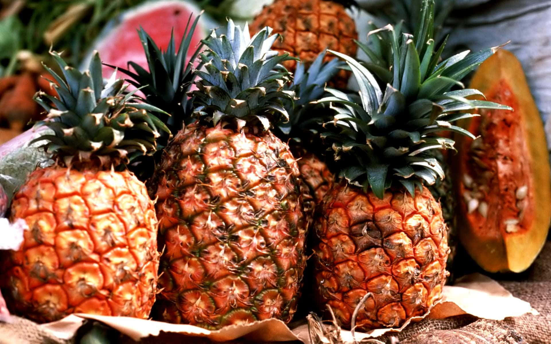 Pineapple 桌布 壁紙 — 成熟的鳳梨帶有尖刺的綠色冠部，旁邊放著切好的西瓜和木瓜，置於質樸的麻布上。