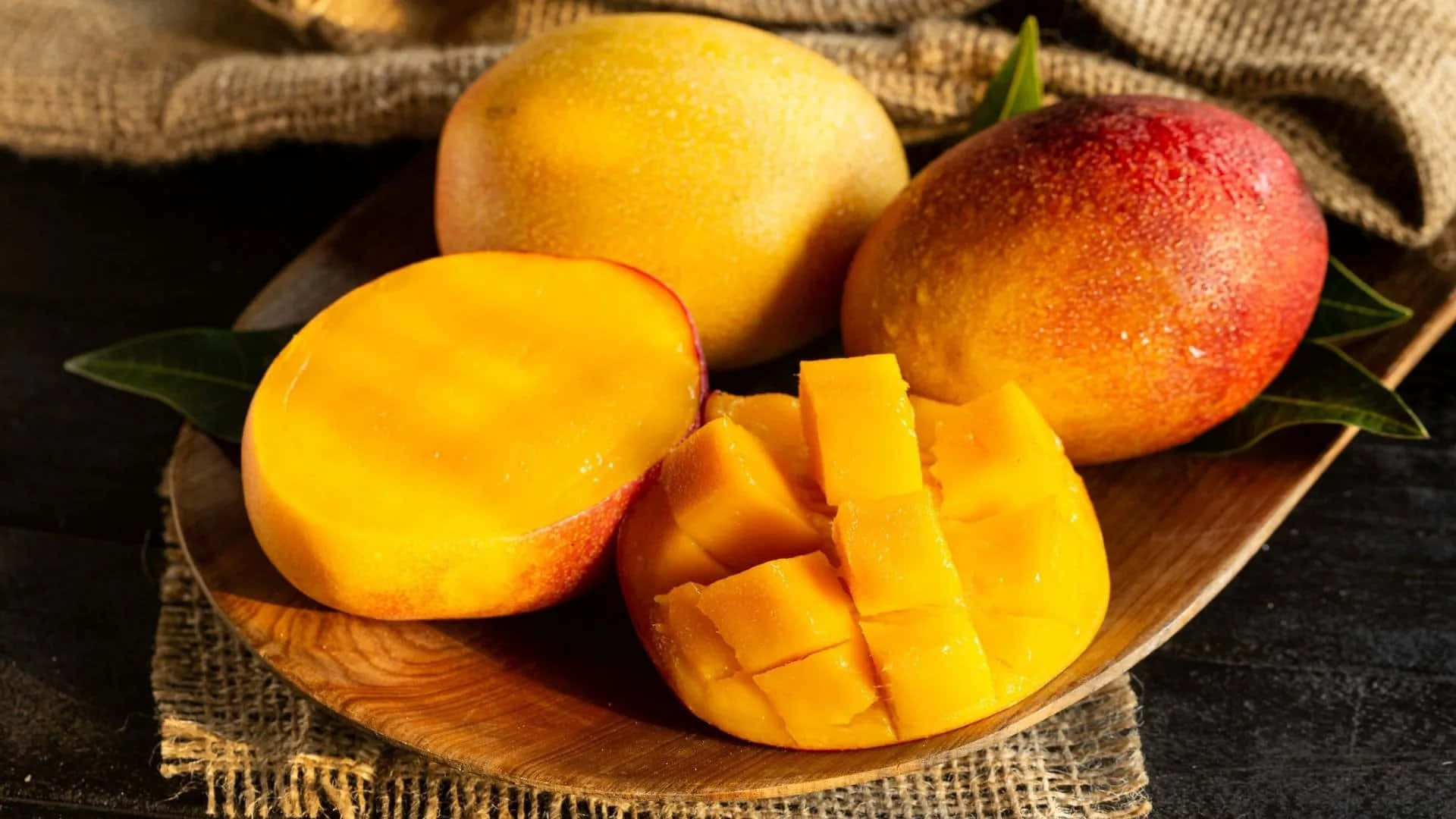 Mango 桌布 壁紙 — 成熟芒果完整和切片在麻布上的木盤上閃閃發光，搭配茂盛的綠葉。