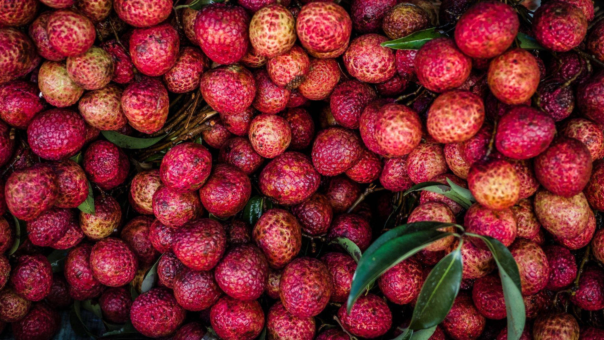 Litchi 桌布 壁紙 — 成熟荔枝的鮮豔特寫，帶有紋理的紅色外皮和散落的綠葉。