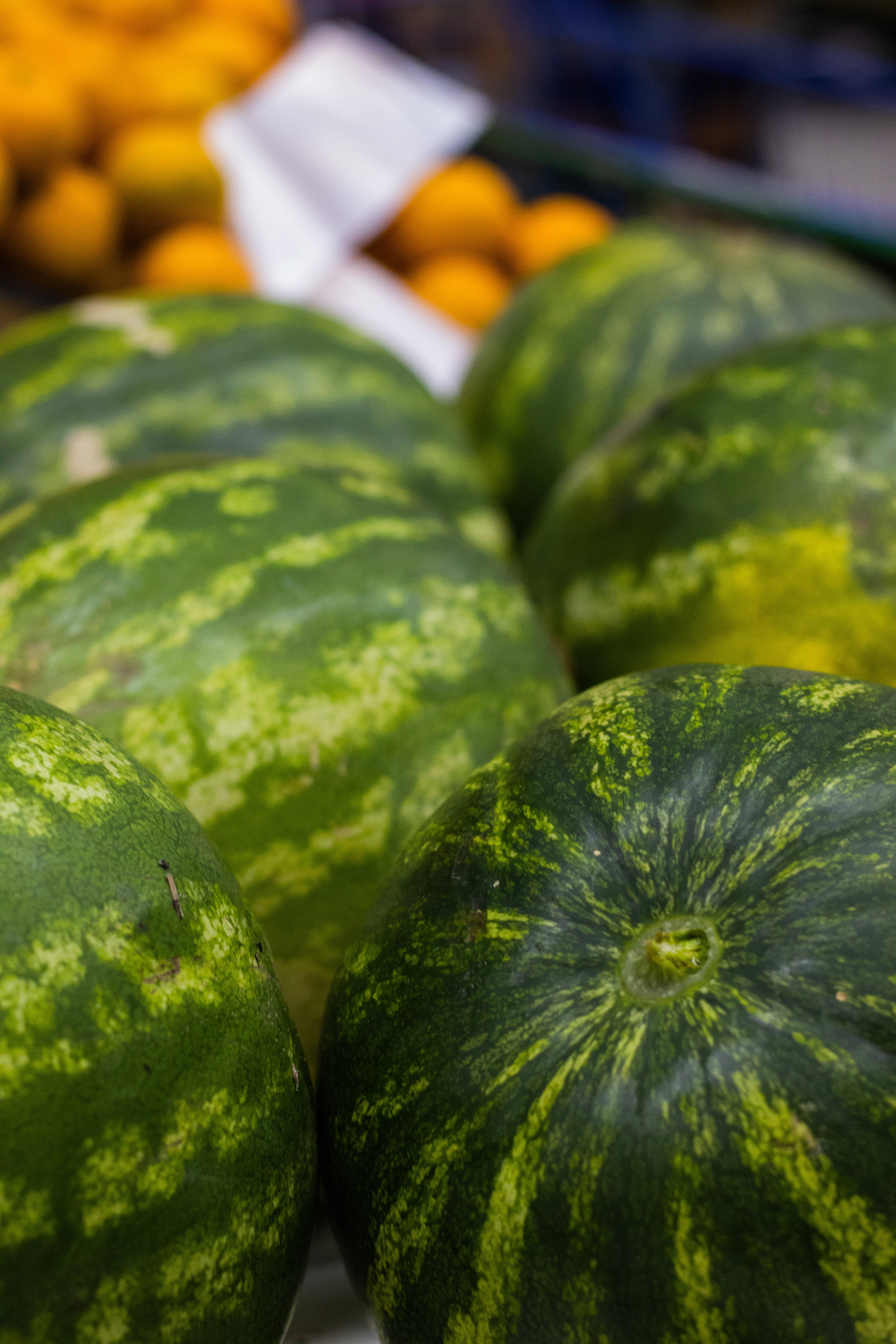 Watermelon 桌布 壁紙 — 成熟西瓜的特寫，綠色條紋外皮堆疊在市場攤位上，背景有模糊的橙子。