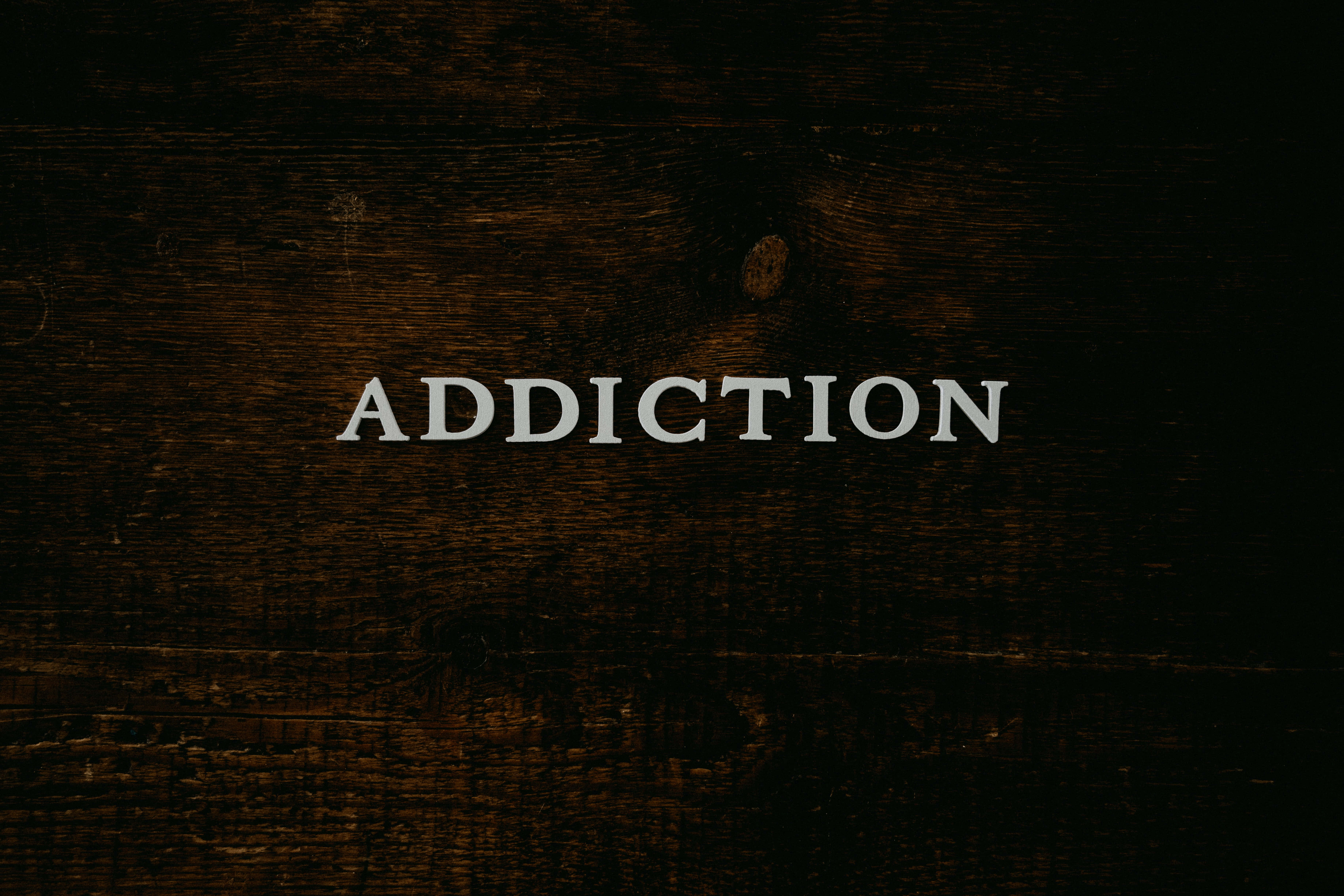 Addiction 桌布 壁紙 — 「成癮」一詞以白色襯線字體居中顯示在帶有可見紋理和節疤的深色木質表面上。