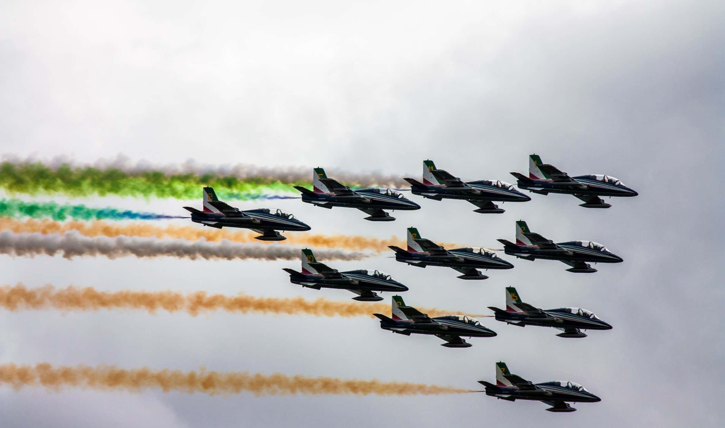 2440x1440 Small Planes 桌布 壁紙 — 戰鬥機以精準編隊劃過多雲的天空，留下鮮豔的彩虹色煙霧軌跡。