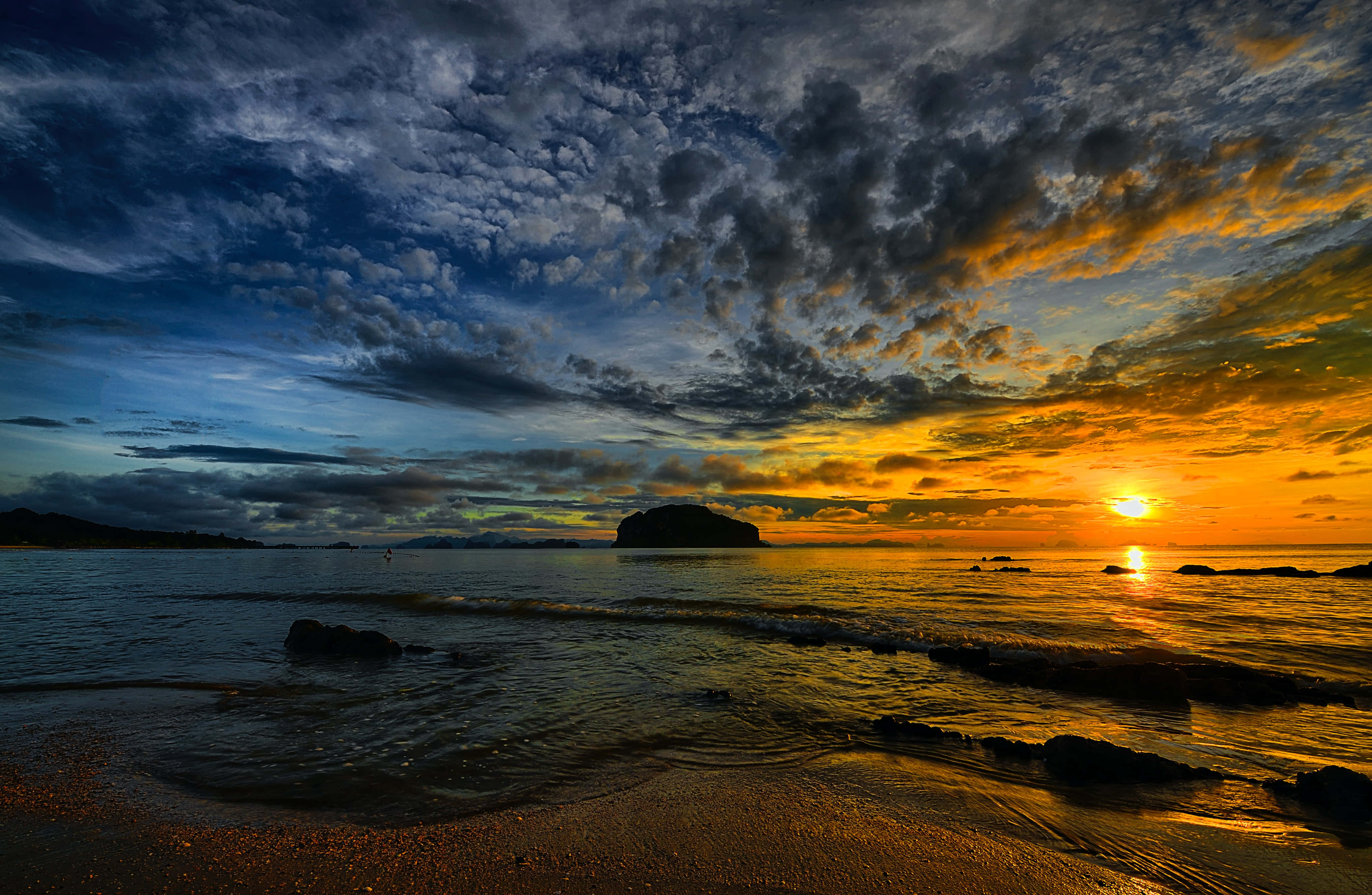 Sunset Beach 桌布 壁紙 — 戲劇性的夕陽將天空染成火熱的橙色和深藍色，海浪拍打岩石海岸，遠處有島嶼的輪廓。