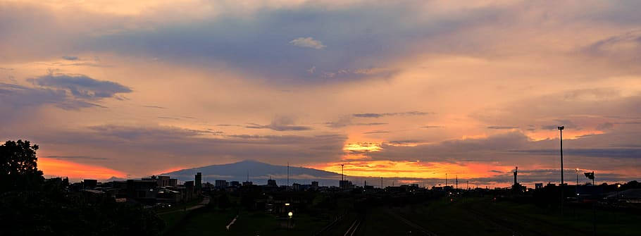 Volcano 桌布 壁紙 — 戲劇性的夕陽將天空染成火熱的橙色和紫色，遠山聳立在剪影般的城市景觀之上。