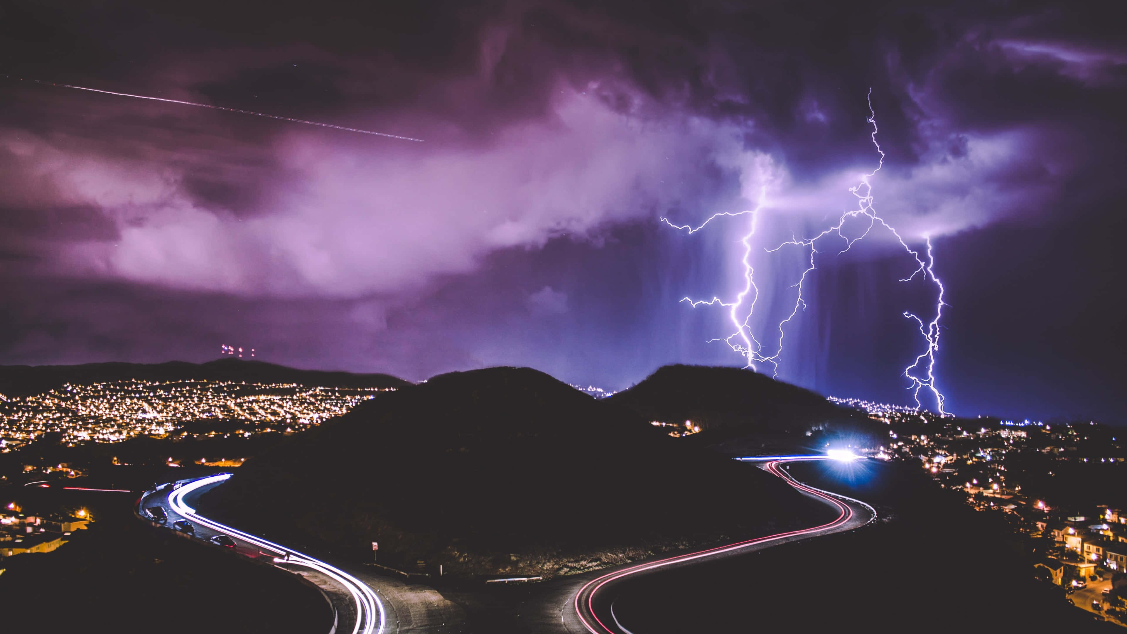 Purple Lightning 桌布 壁紙 — 戲劇性的夜景，鮮豔的紫色閃電照亮了蜿蜒道路和下方閃爍城市燈光之上的暴風雨天空。