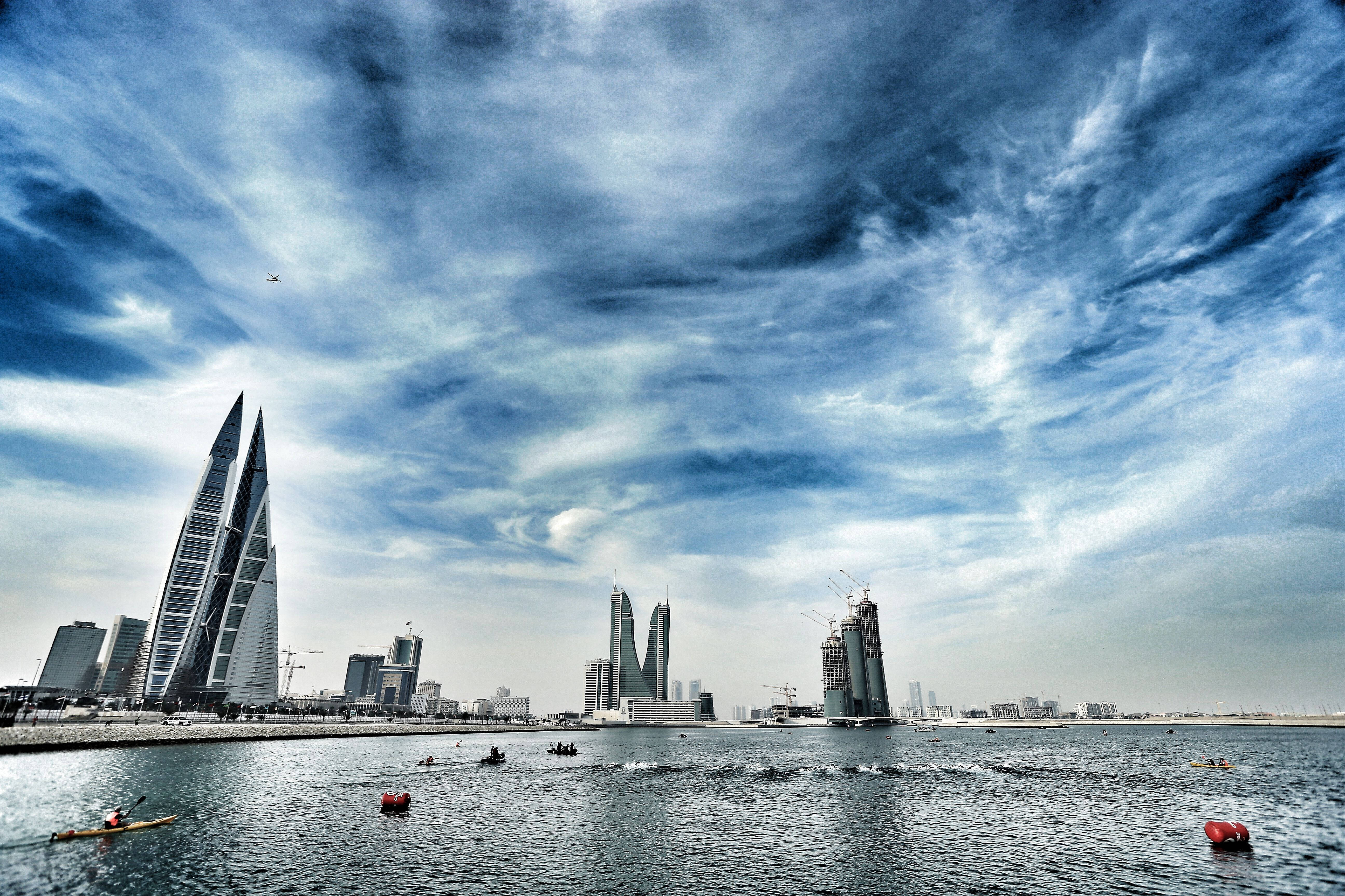 Bahrain 桌布 壁紙 — 戲劇性的天空，漩渦狀的雲層籠罩著現代摩天大樓和寧靜水面上的皮划艇愛好者。