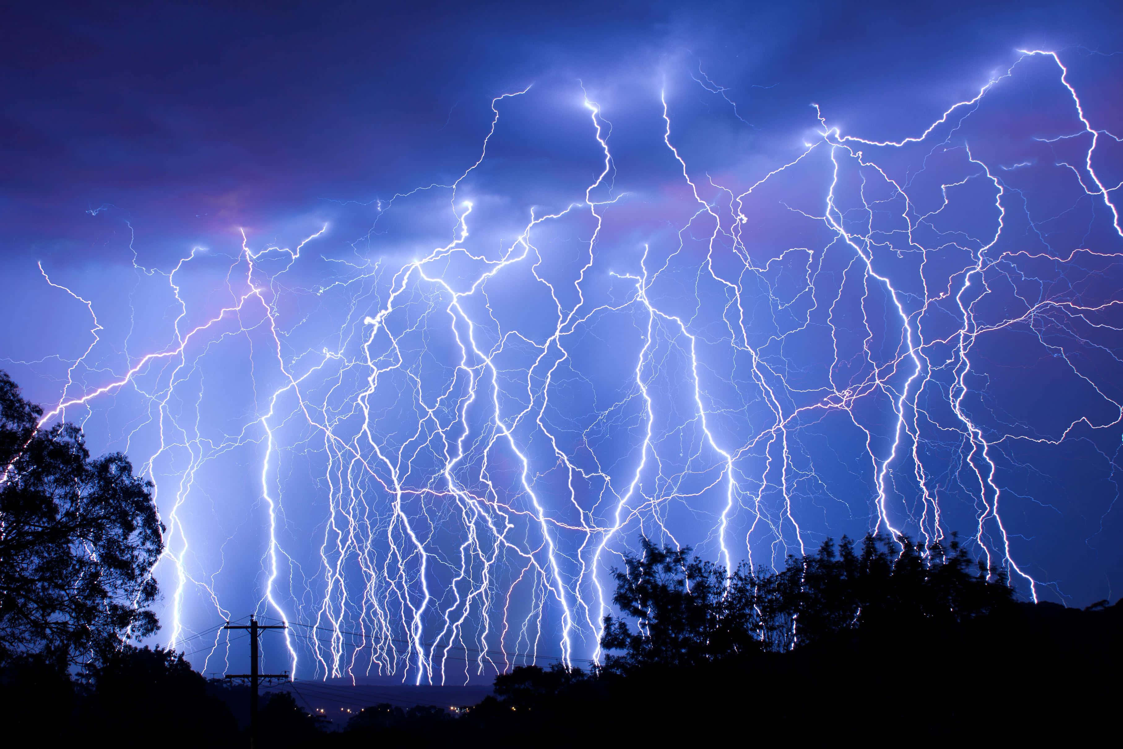 thunder 桌布 壁紙 — 戲劇性的天空充滿強烈的白色和紫色閃電，照亮了黑暗的雲層，映襯出樹木和電線杆的剪影。